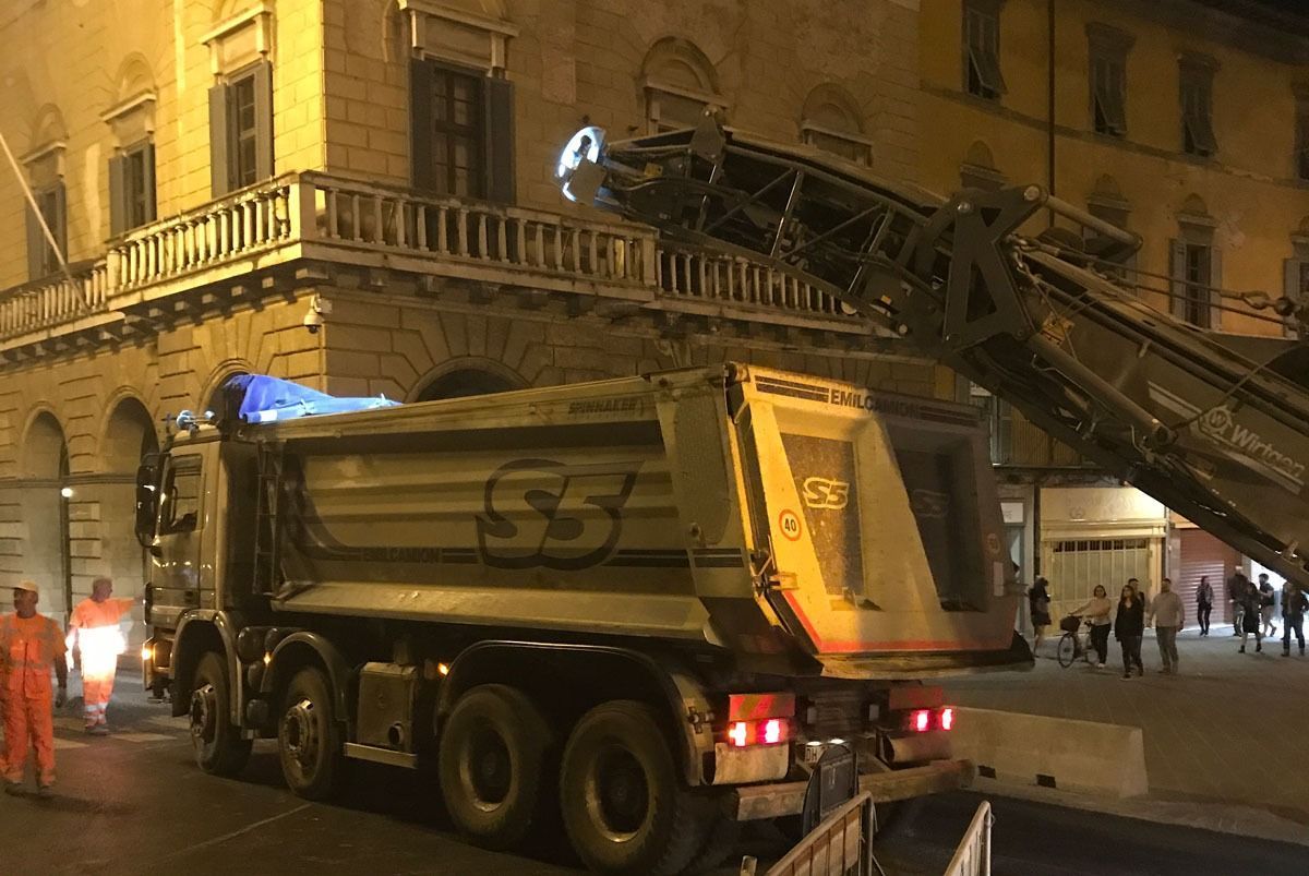 Un camion con cassone ribaltabile che riceve asfalto da una fresatrice su una strada cittadina di notte. Operai con gilet arancioni nelle vicinanze.