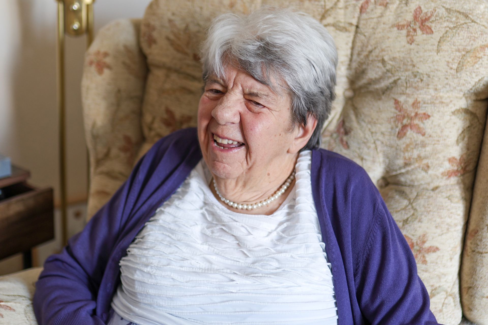 An elderly person with gray hair, wearing a white shirt, pearl necklace, and purple cardigan, smiles while seated.