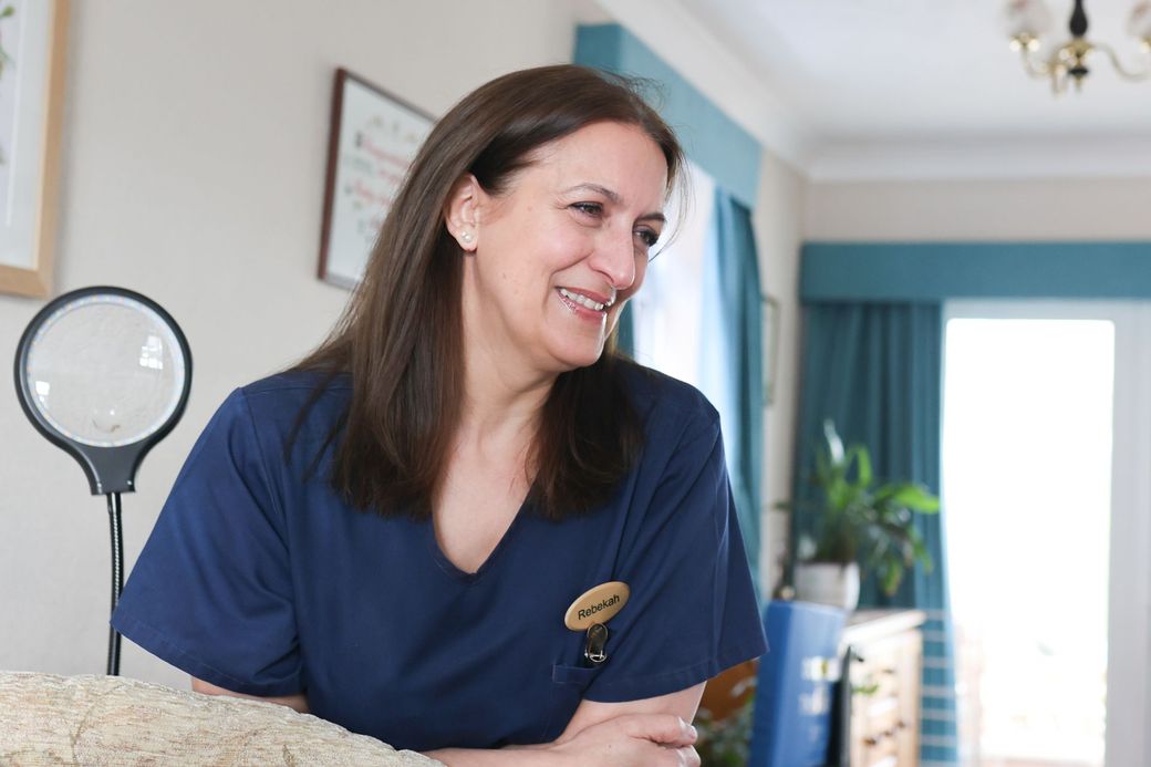A person in a blue scrub top with a name tag, smiling while standing in a bright office with a magnifying lamp.