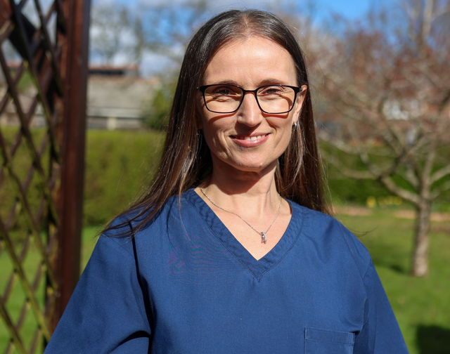 A professional smiling in blue scrubs and glasses, posing outdoors in front of a garden.