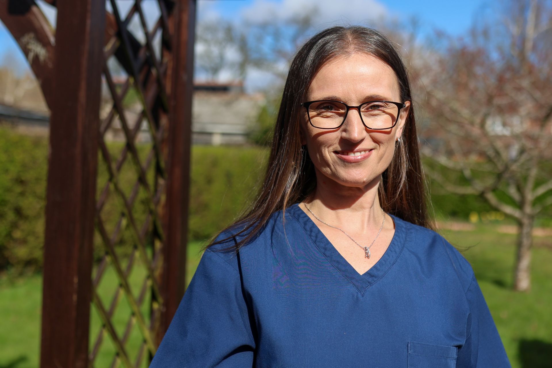 A person with long brown hair and glasses wearing a blue scrub top, standing outdoors in a garden setting.