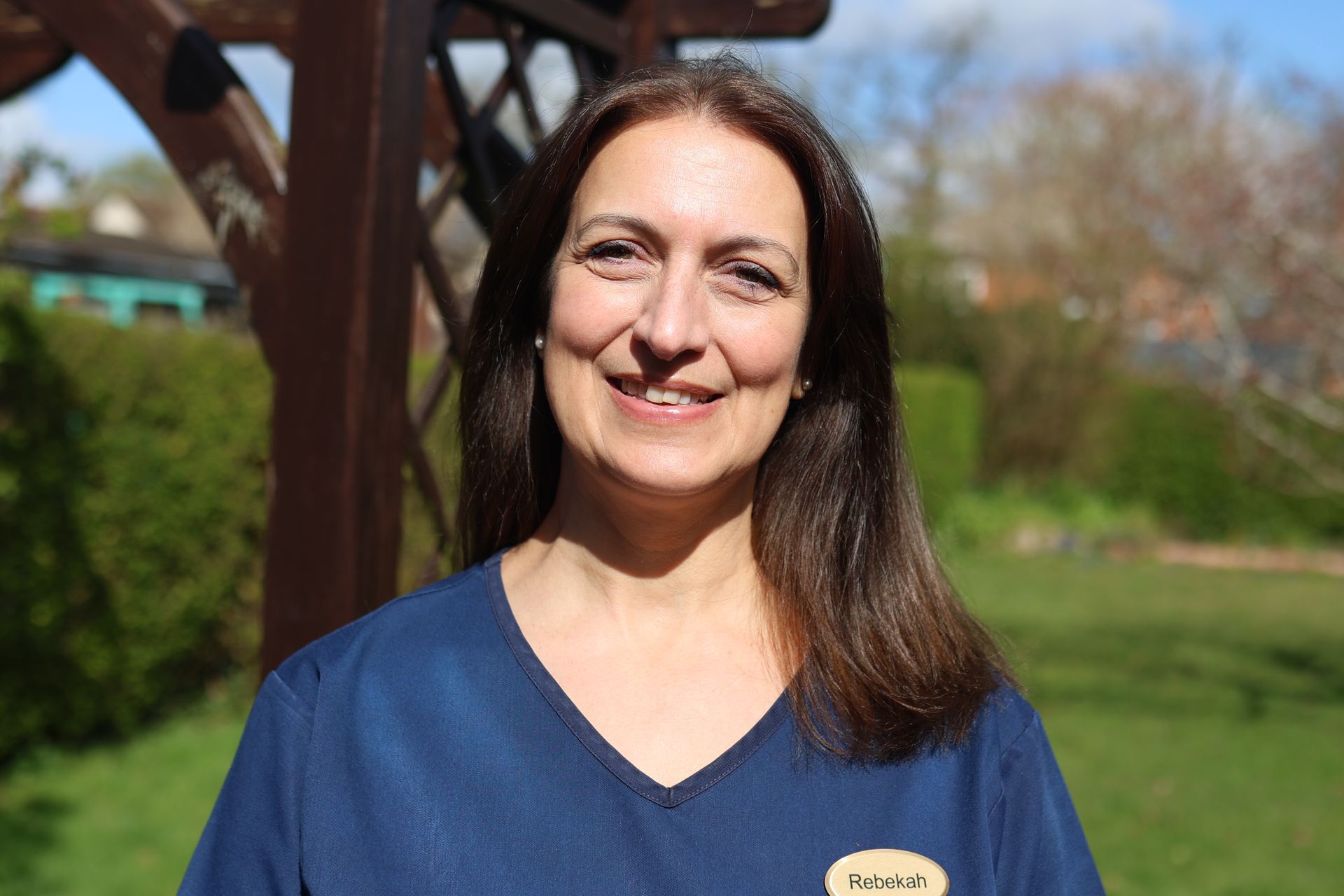 A smiling person wearing a blue scrub top with a name tag, standing outdoors in front of a wooden pergola and greenery.