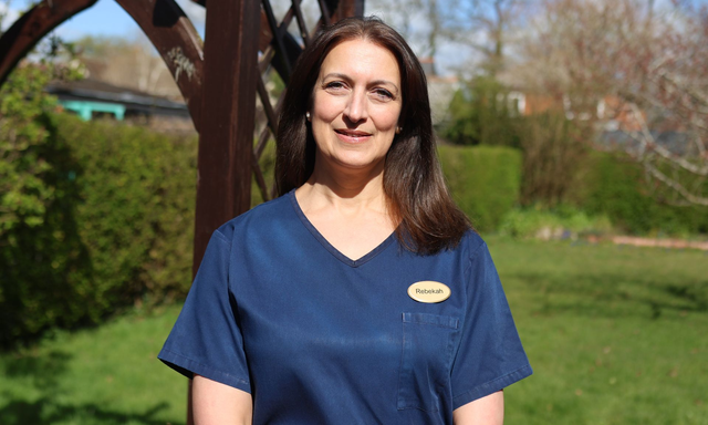 A person in a dark blue V-neck uniform with a name tag stands outdoors in front of a garden archway.