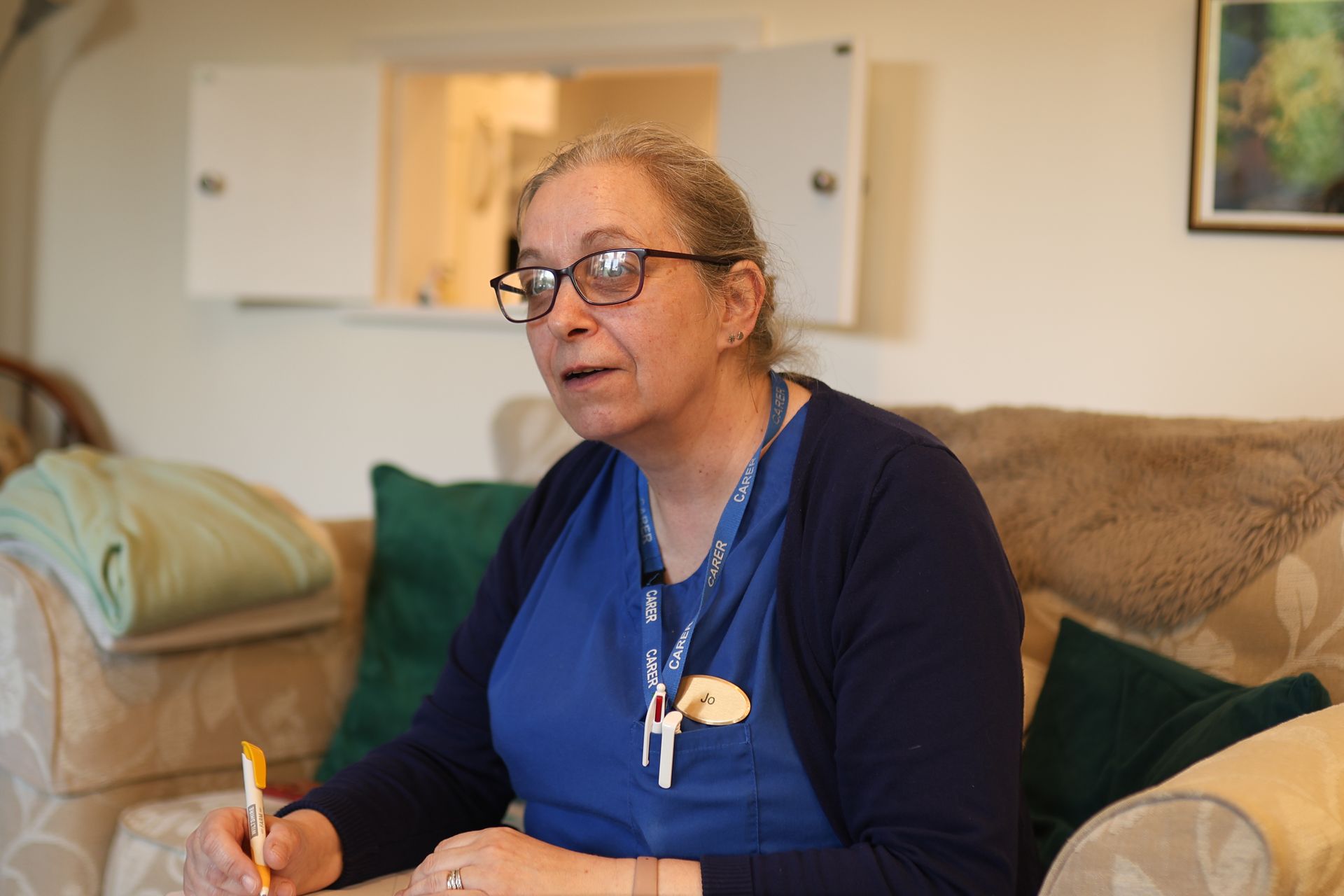 A person wearing a blue uniform and an NHS lanyard sits on a couch in a living room, holding a pen and speaking.