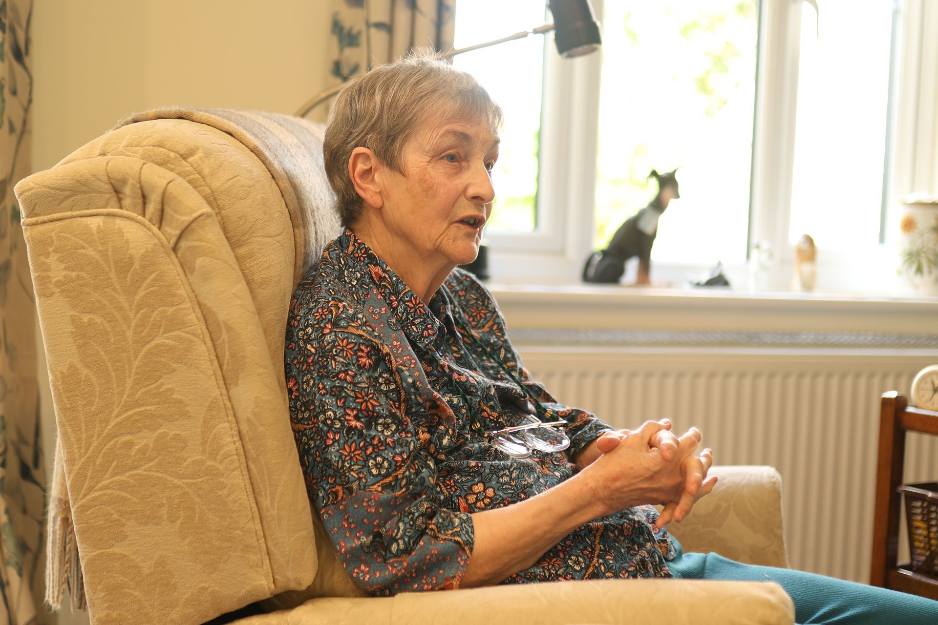 A person with short hair sits in a light-colored armchair, hands clasped, looking slightly to the side in a home setting.