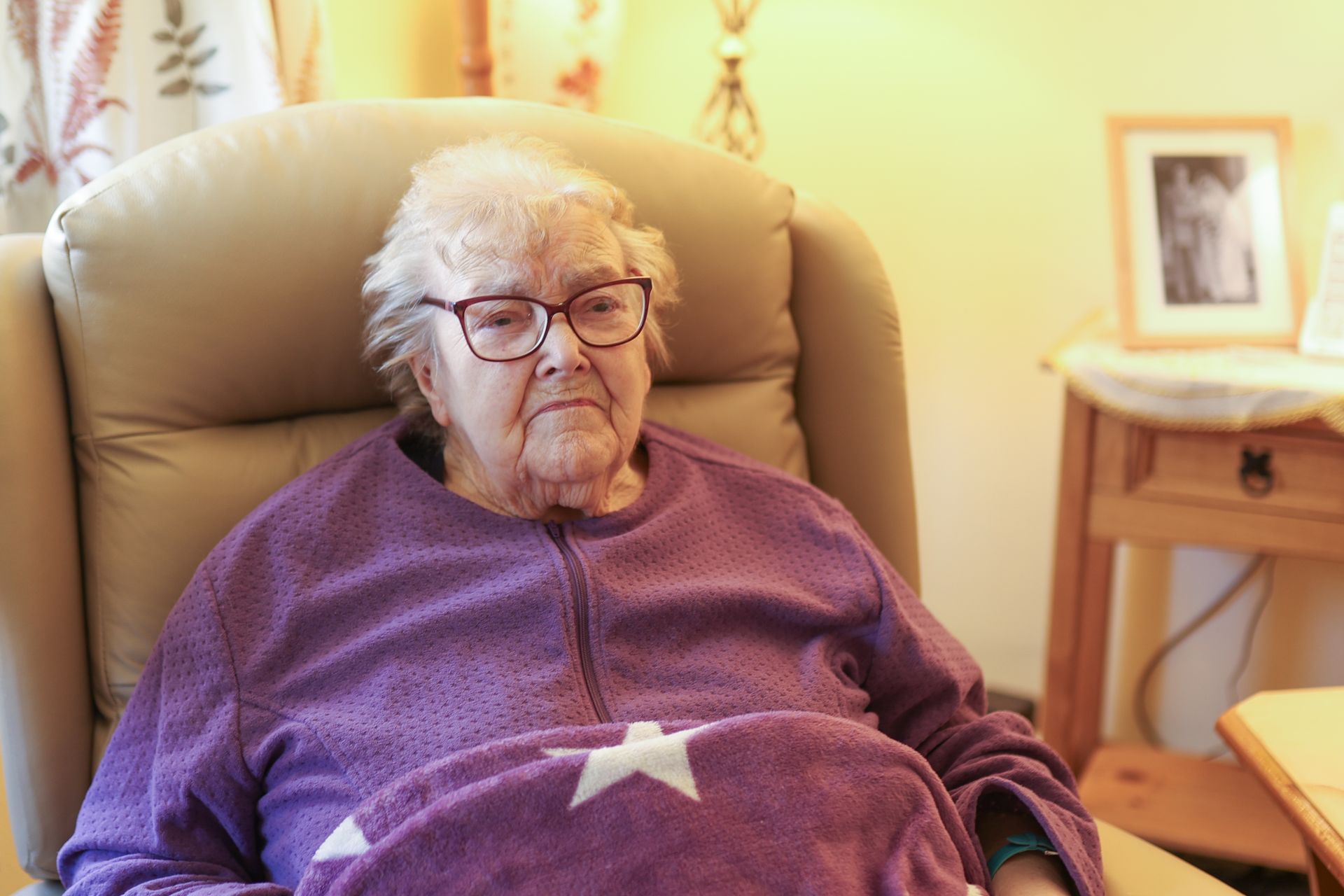 An older person wearing glasses and a purple star-patterned sweater sits in a tan armchair.