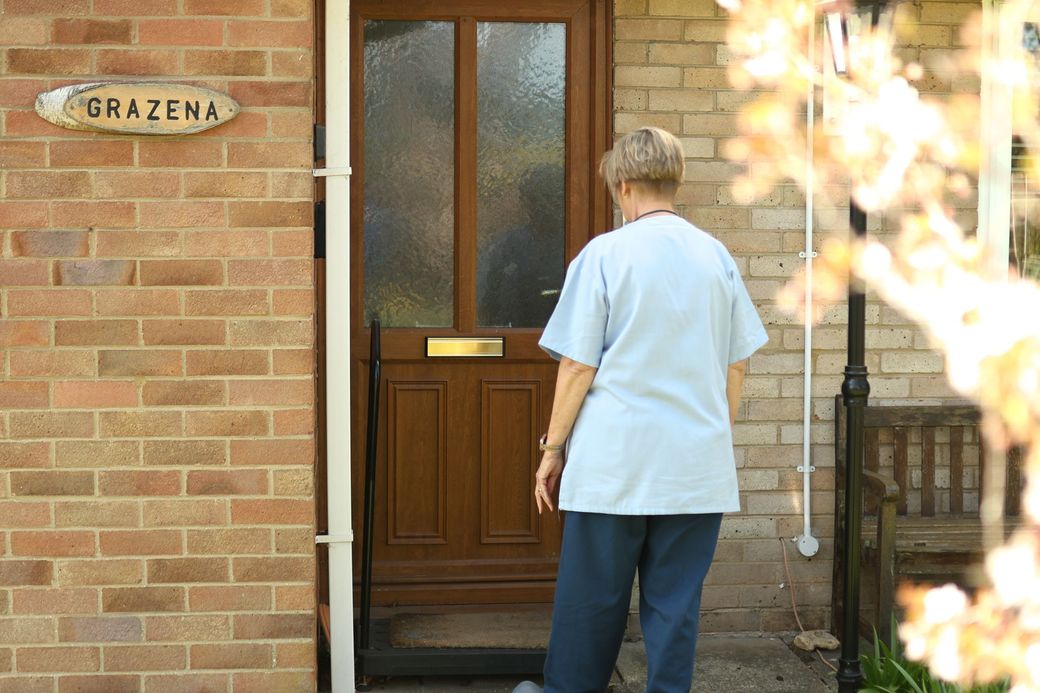 A person in a light blue shirt and dark pants stands before a brown front door with a nameplate labeled “GRAZENA.”