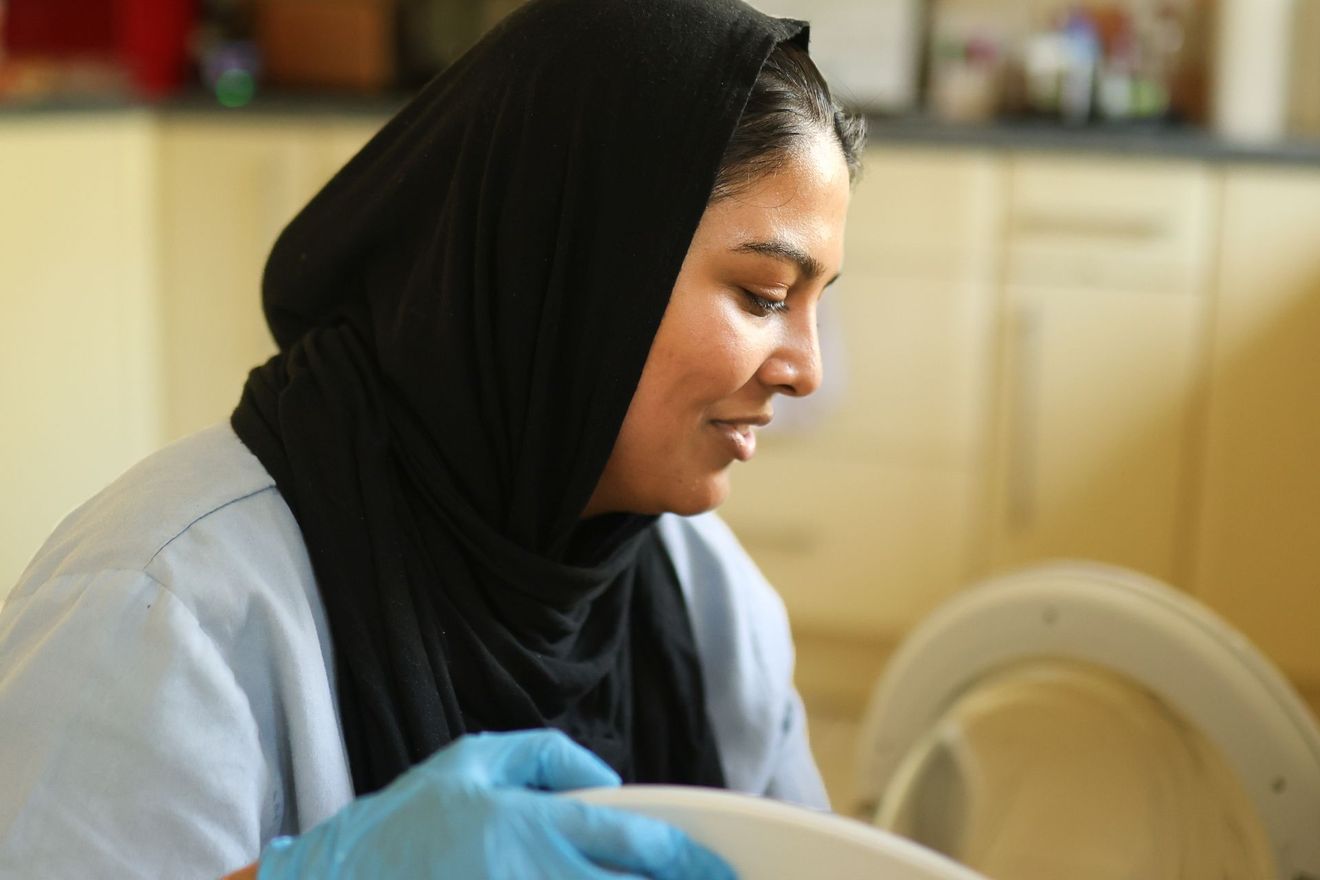 A person in a black headscarf and light blue scrubs wearing blue medical gloves while working in a kitchen.