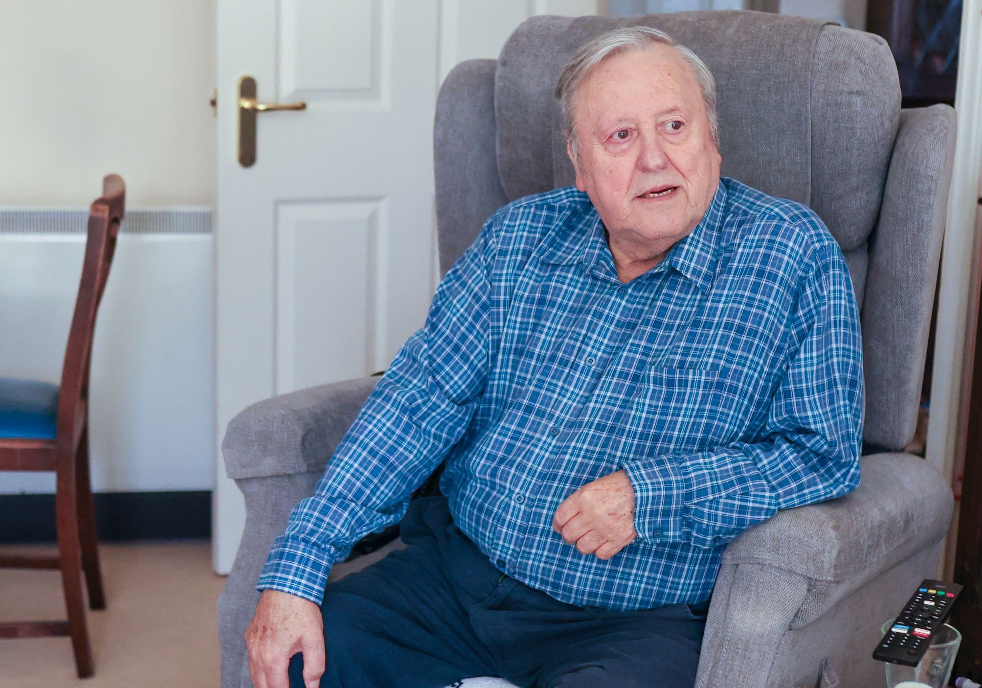 An individual wearing a blue patterned long-sleeved shirt sits in a grey armchair in an indoor setting.
