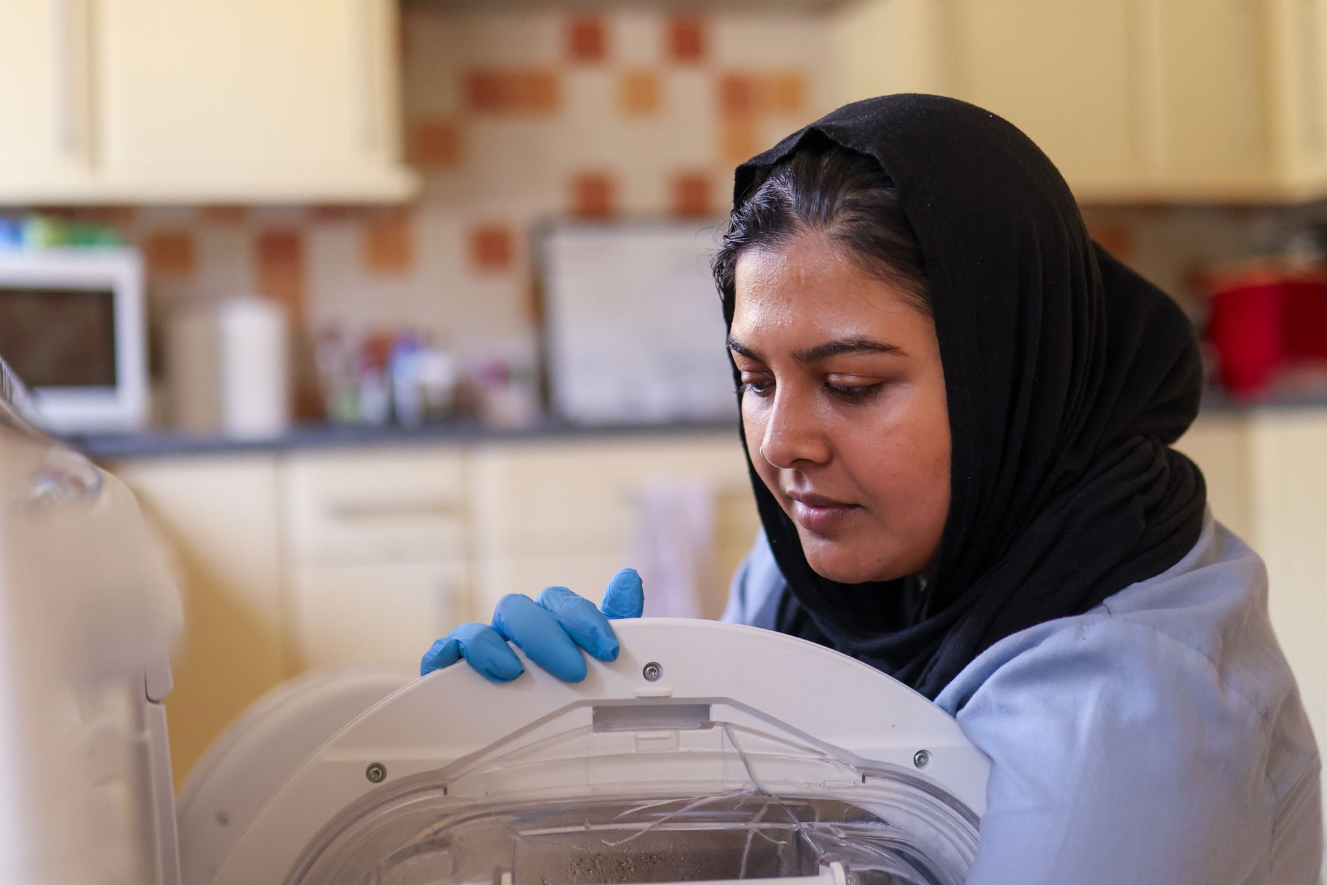 A person wearing a hijab and blue gloves opens the door of a washing machine in a kitchen.