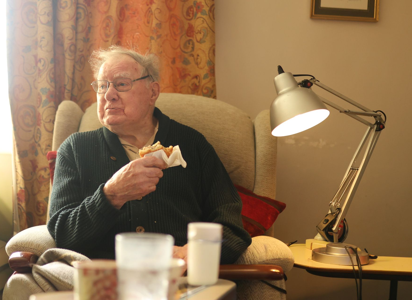 A person sits in a beige armchair, eating while looking to the side next to a desk lamp.