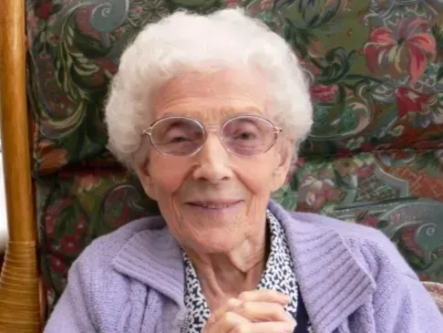 An elderly person with white hair and glasses smiles while wearing a lavender cardigan over a patterned shirt.