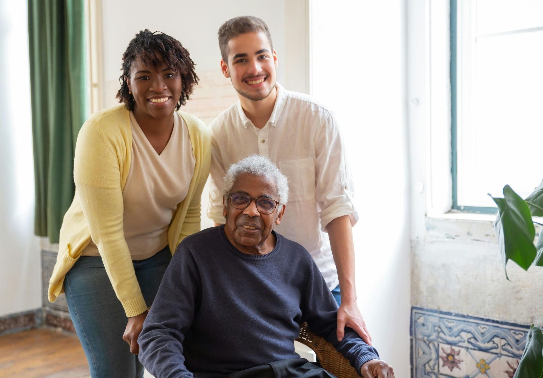 Two people stand behind a smiling person sitting in a chair, all positioned near a window in a brightly lit room.