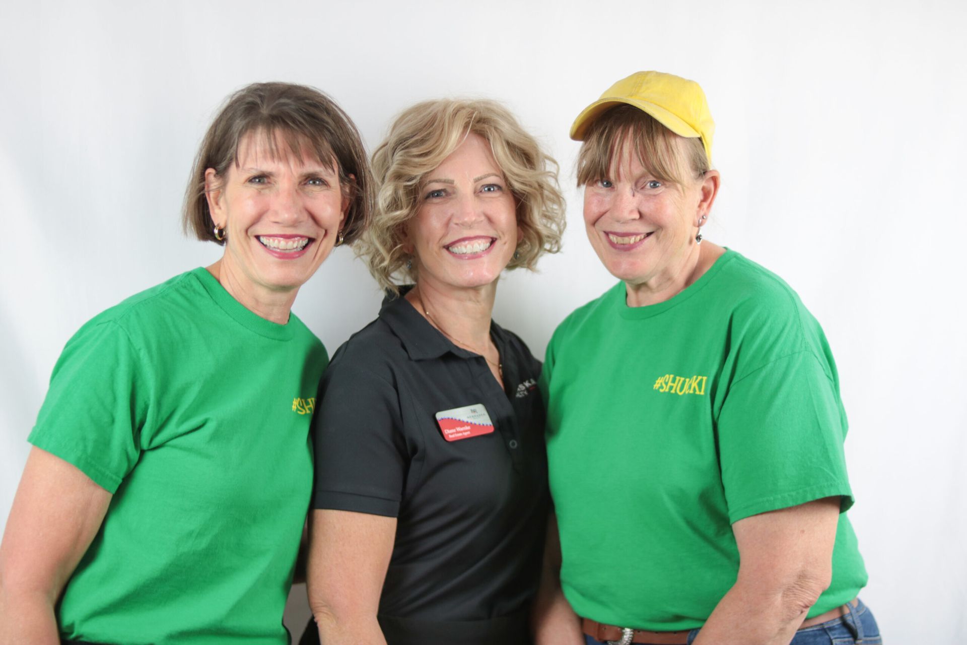 Three women smiling, two in green shirts, one in a black shirt, posing in front of a white backdrop.