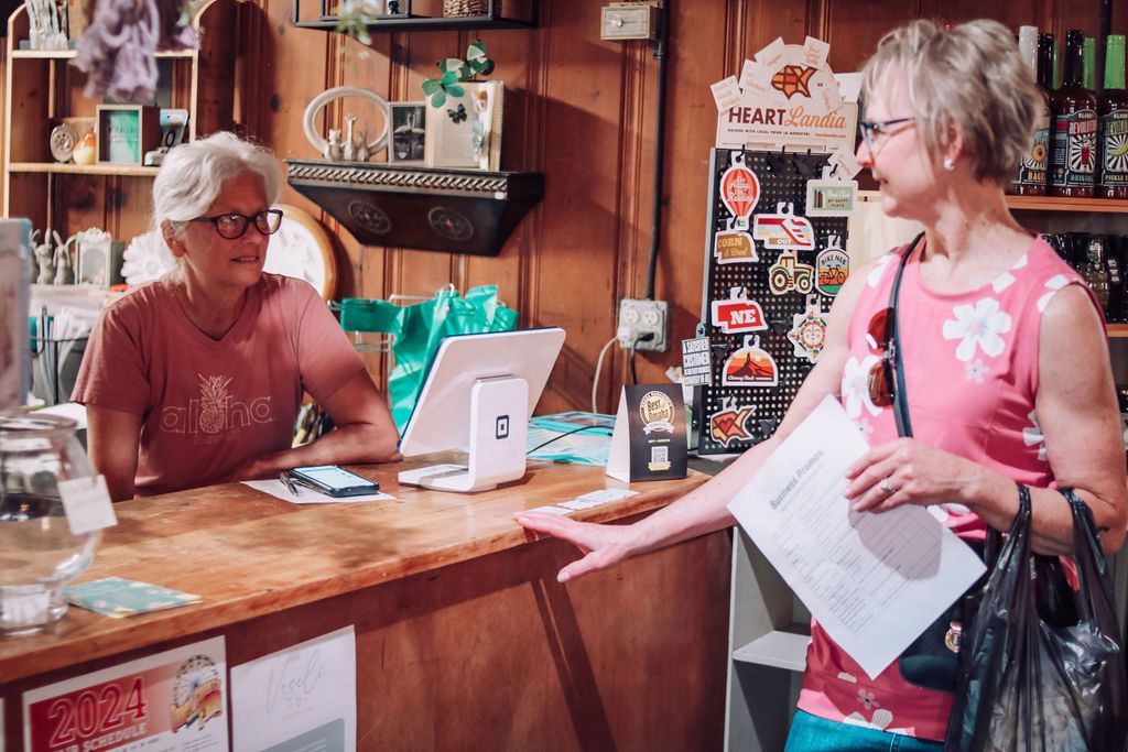 Two women at a shop counter. One gestures, holding papers. The other sits, appearing to work. Interior shot.
