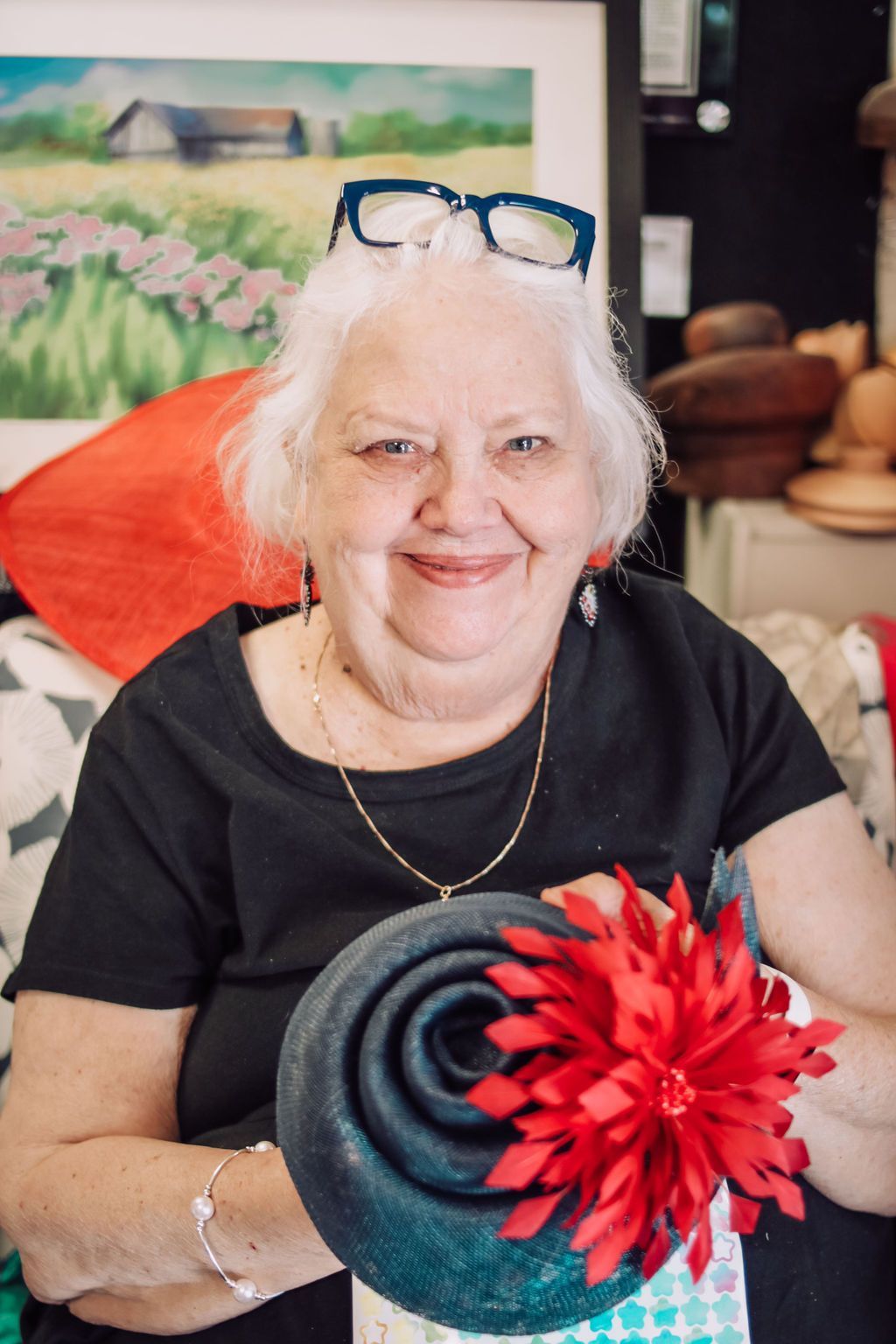 Woman holding a dark blue hat with a red flower. She smiles, wearing glasses, and is in a hat shop.