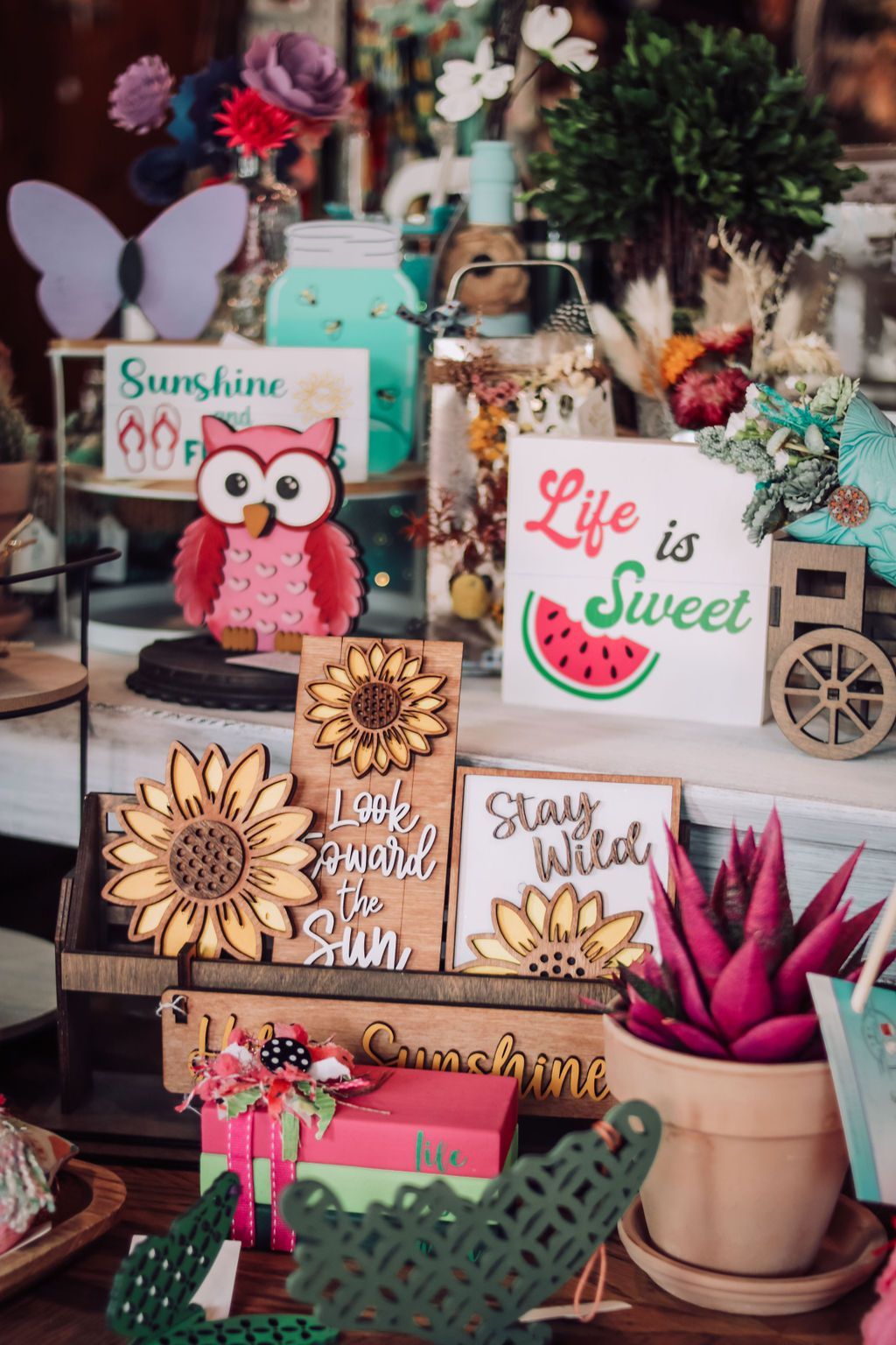 Display of decorative wooden signs, an owl, and other colorful floral and summer-themed crafts.