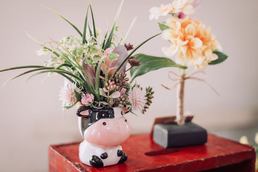 Cow-shaped vase with colorful flowers sits atop a red box. Another flower arrangement is in the background.