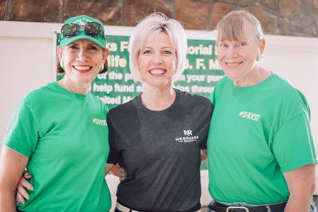 Three women smiling: Two wearing green shirts stand with a woman in black. Sign in background.