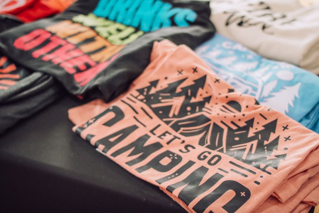 T-shirts with camping and colorful slogans for sale, on a black table.