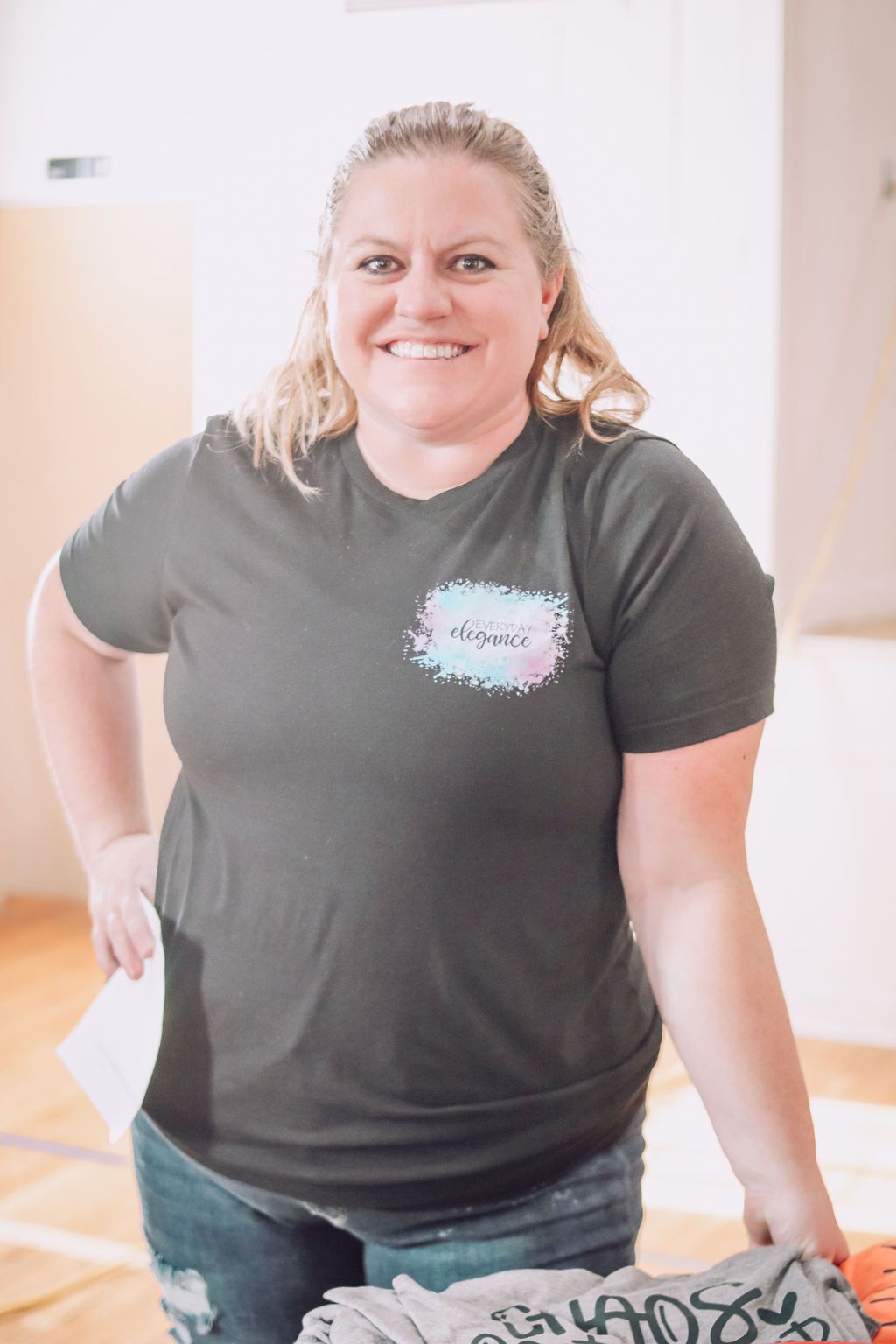 Woman in a dark t-shirt and jeans smiles, holding paper, in a bright room.
