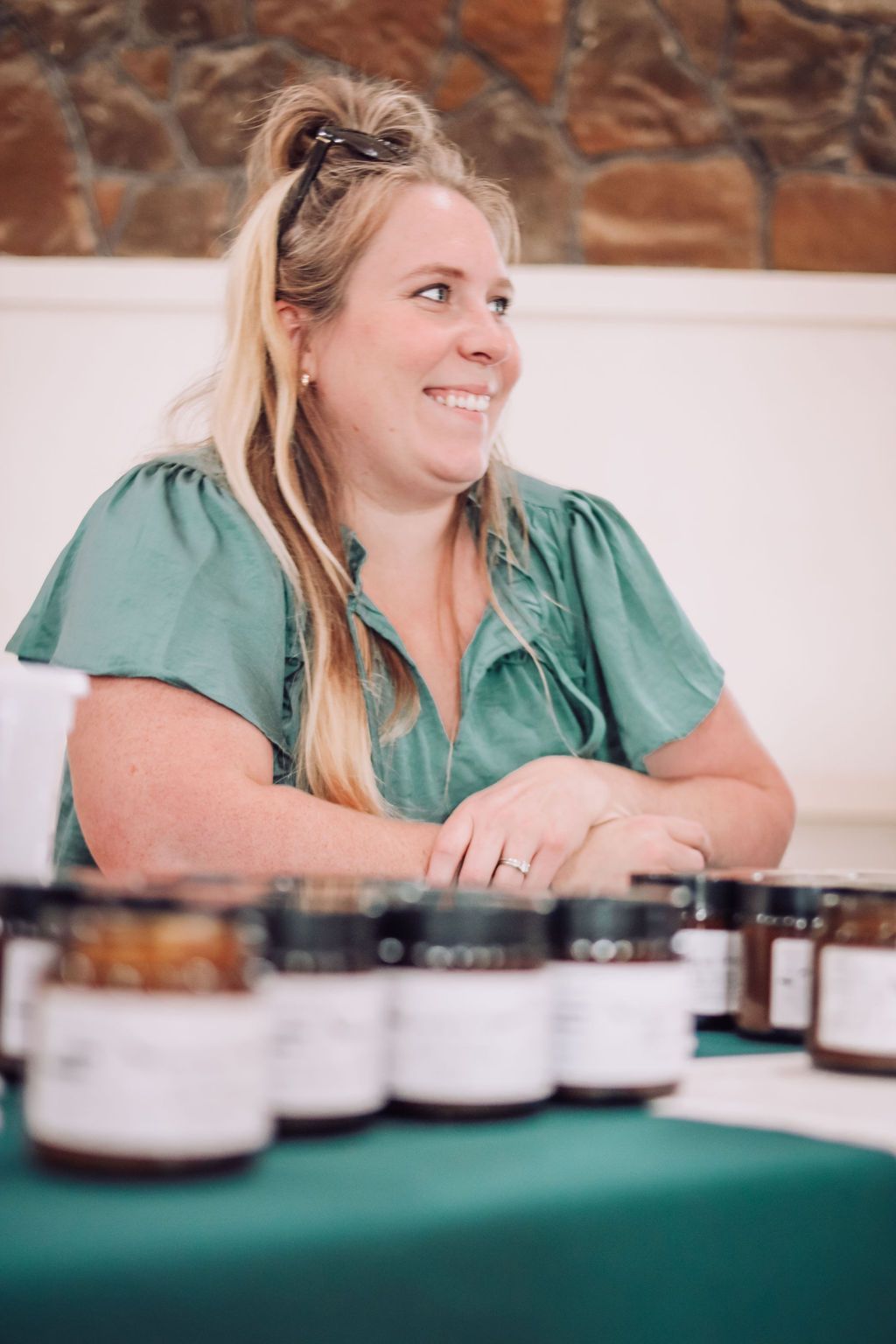 Woman smiling, sitting at a table with jars of product. She wears a green blouse.