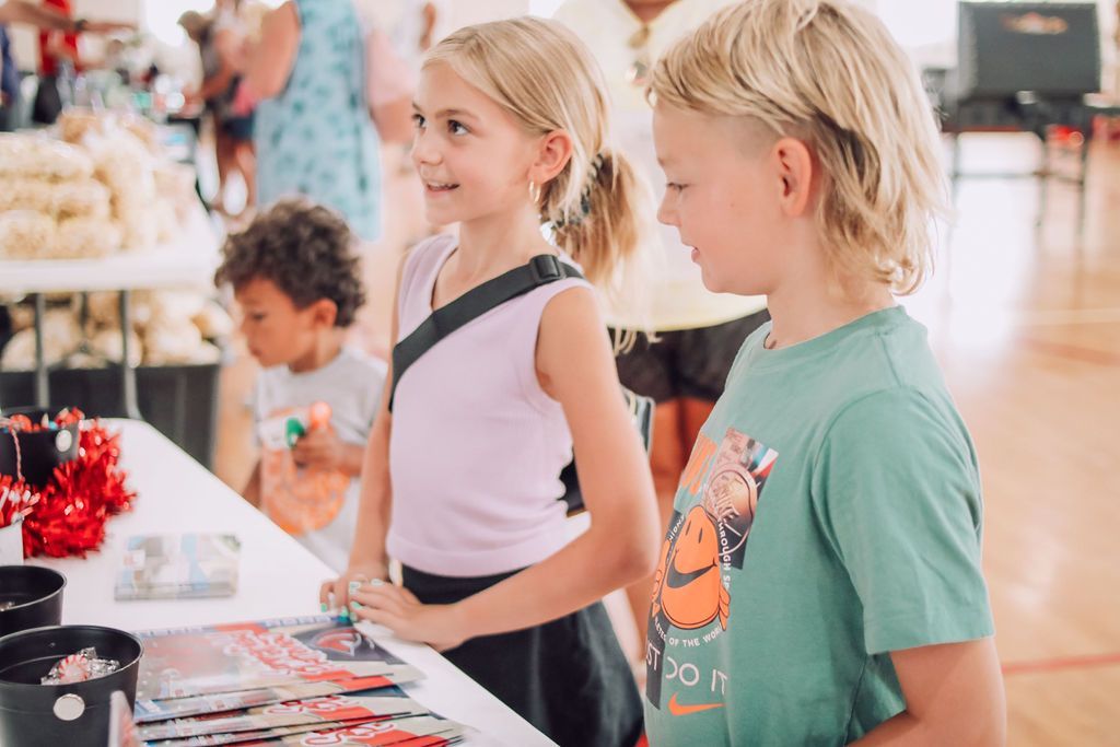 Two children looking at a table with items, others in the background at an event.