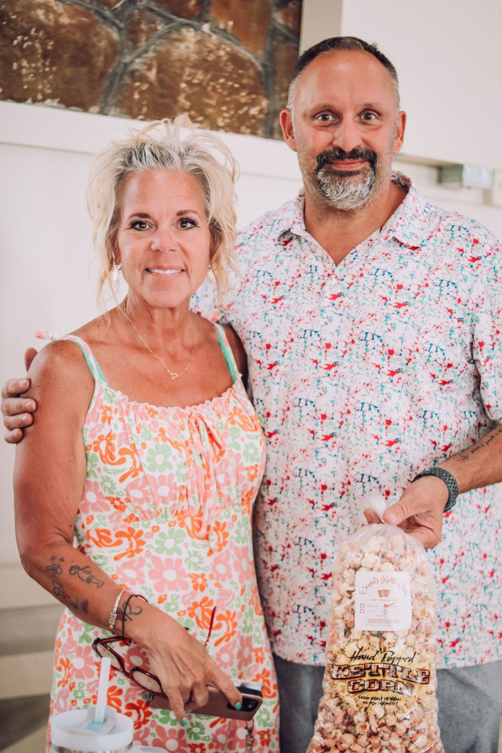 Woman and man holding popcorn bag, smiling. Man's arm around woman. They are indoors.