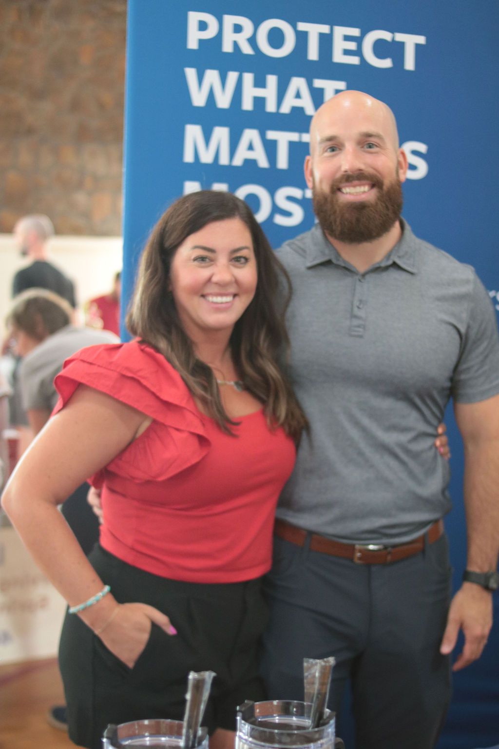 Woman in red top and man with beard pose in front of a blue sign.