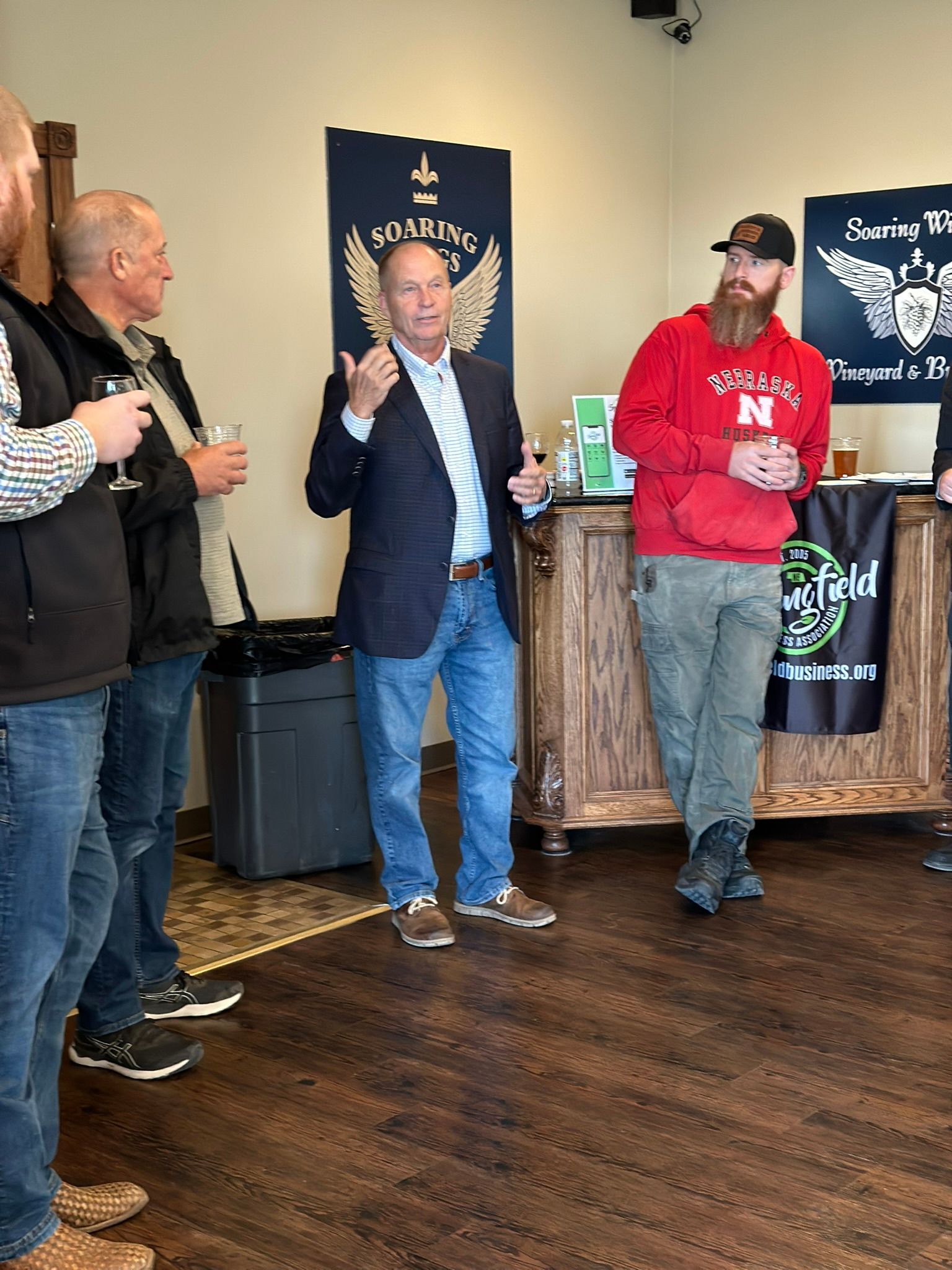 Men gathered indoors; one speaks with hand gestures. Another wears a red sweatshirt.  Logo visible: 