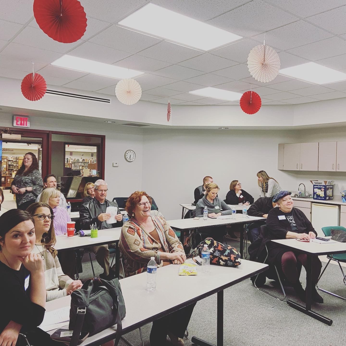 Classroom with people seated at desks, some smiling. Red and white paper decorations hang from the ceiling.