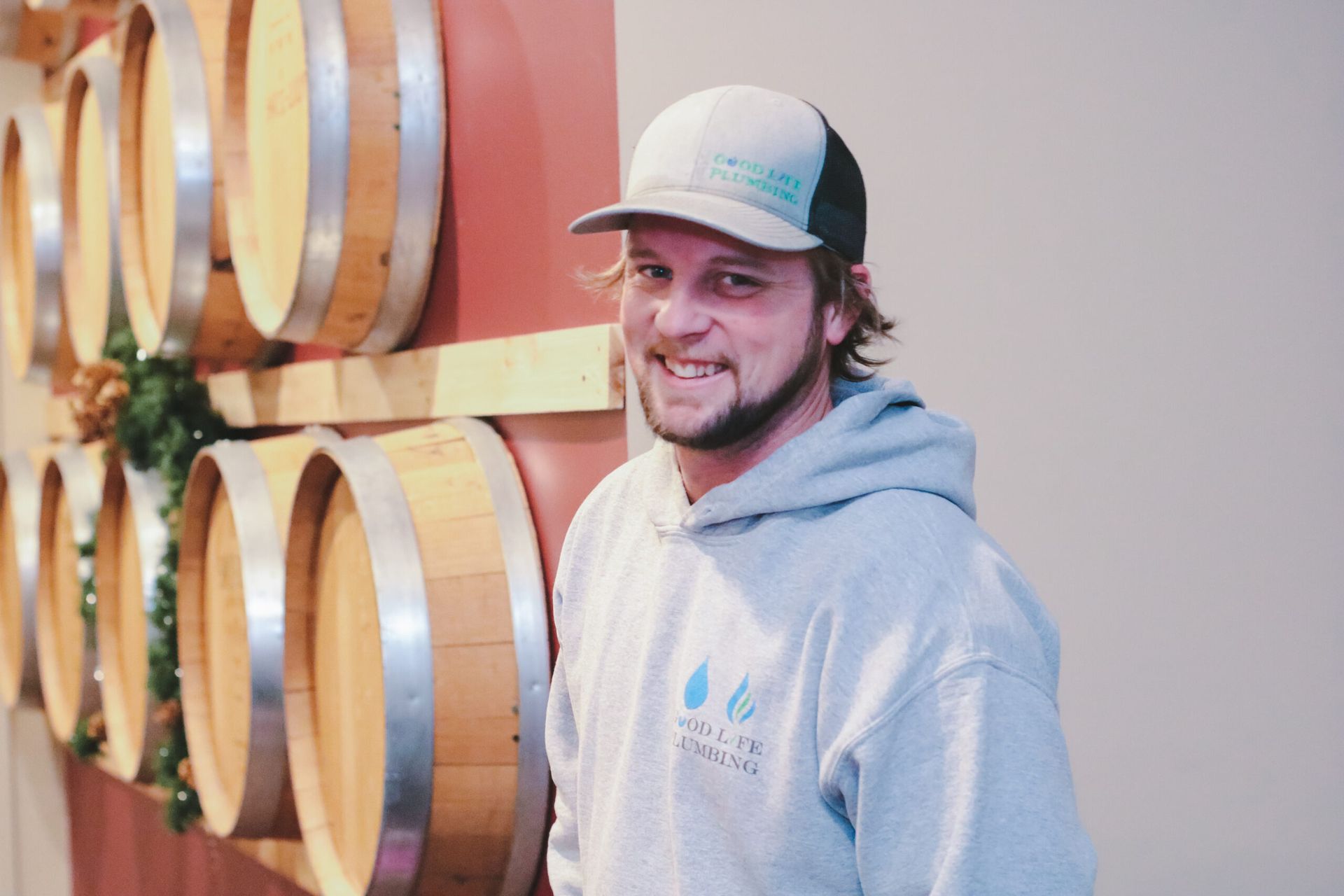 Man wearing a cap and gray hoodie smiles in front of wine barrels.