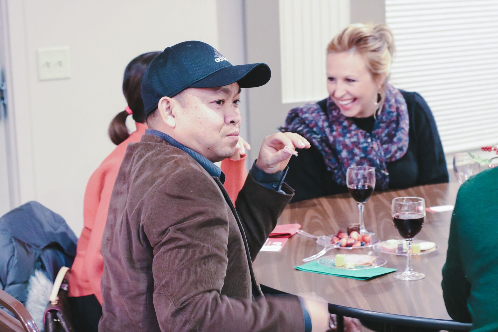 Man in cap gestures while talking, woman laughs. Table with wine, food. Indoors, bright.