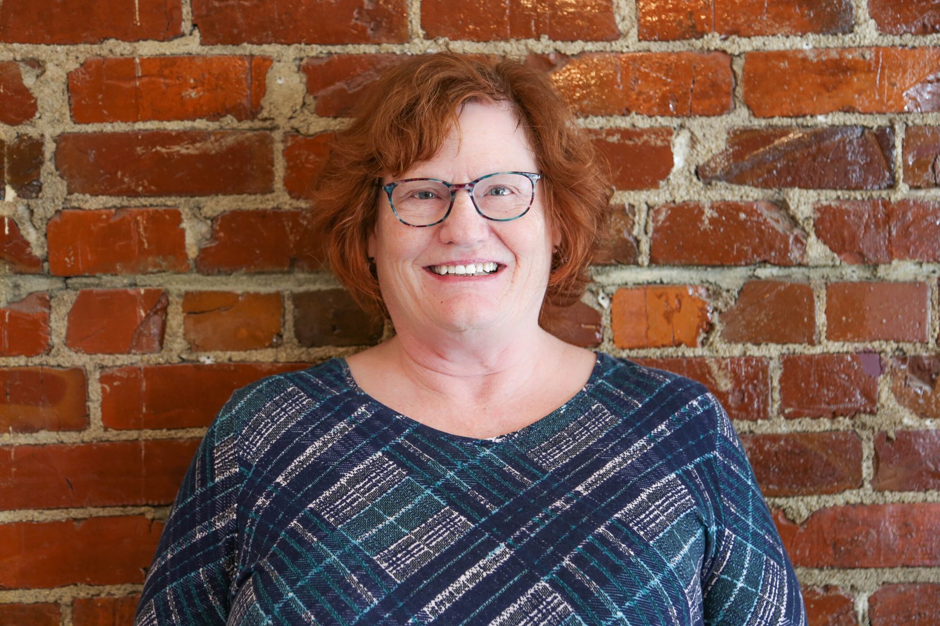 Woman with red hair, glasses, smiling in front of a brick wall, wearing a blue patterned top.