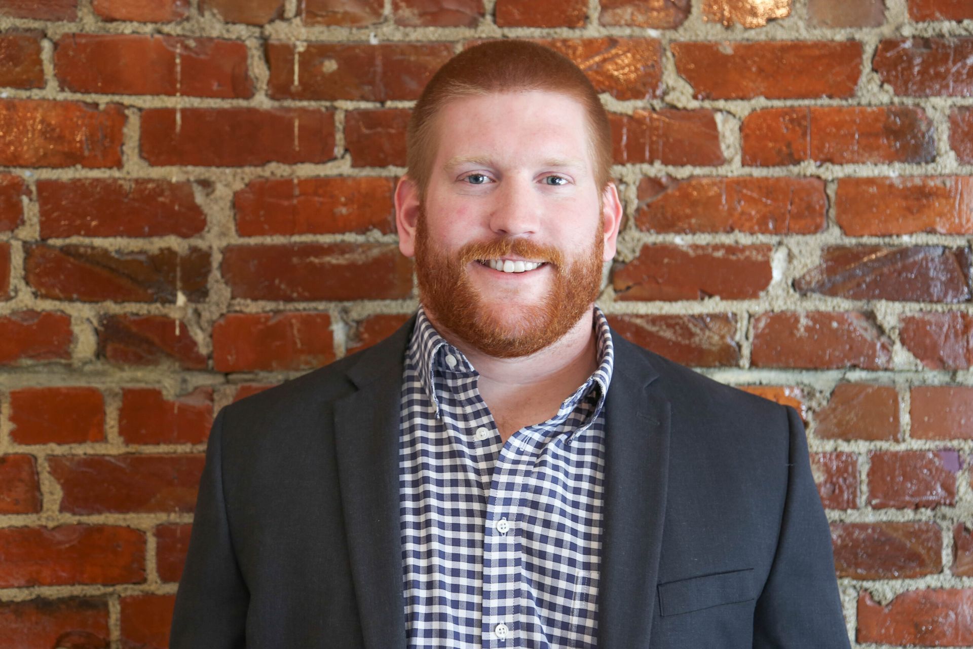 Man with red hair and beard smiles in front of a brick wall, wearing a plaid shirt and jacket.