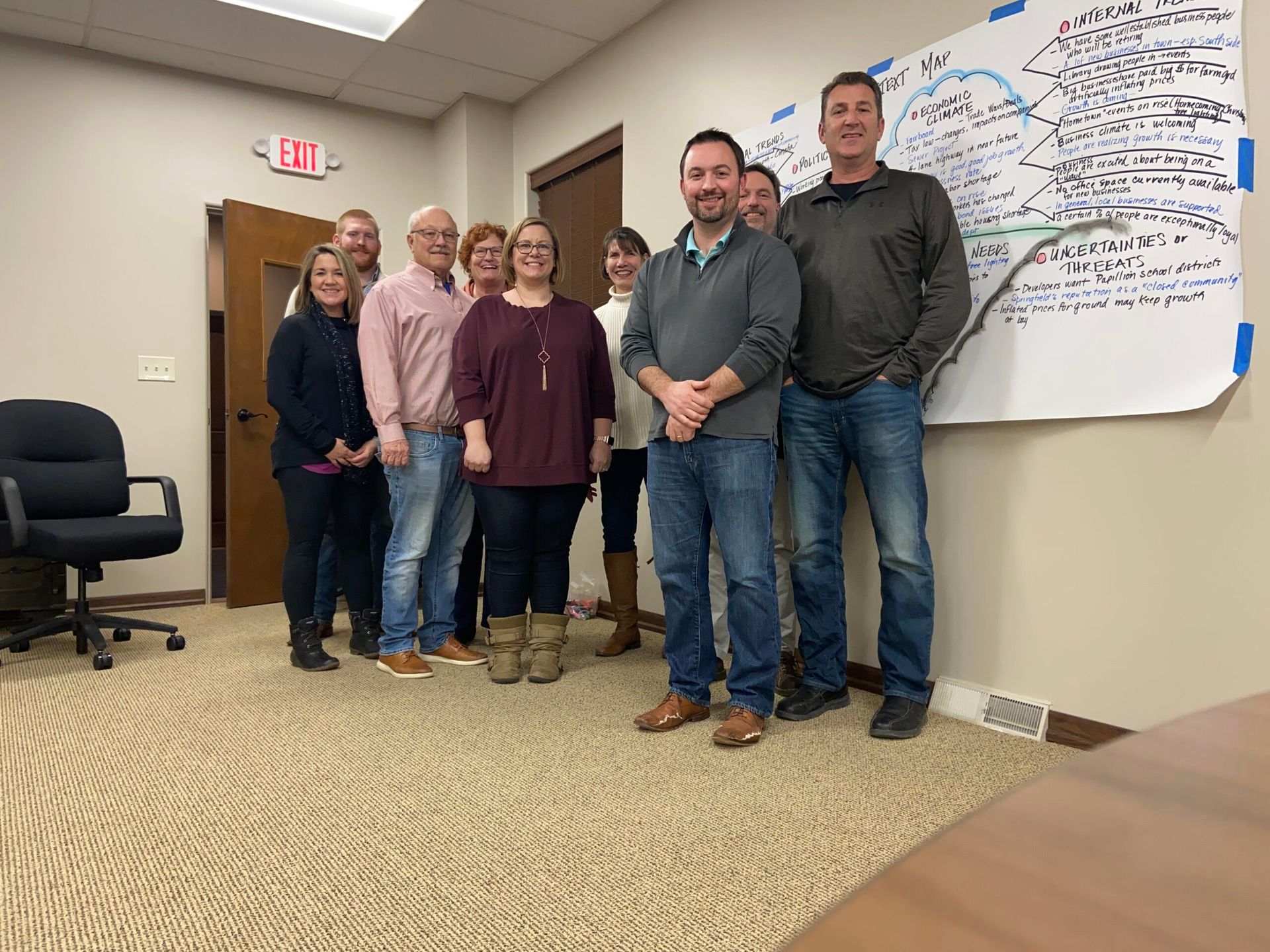 Group of people standing in a room near a whiteboard.  Some are smiling.  Office setting.