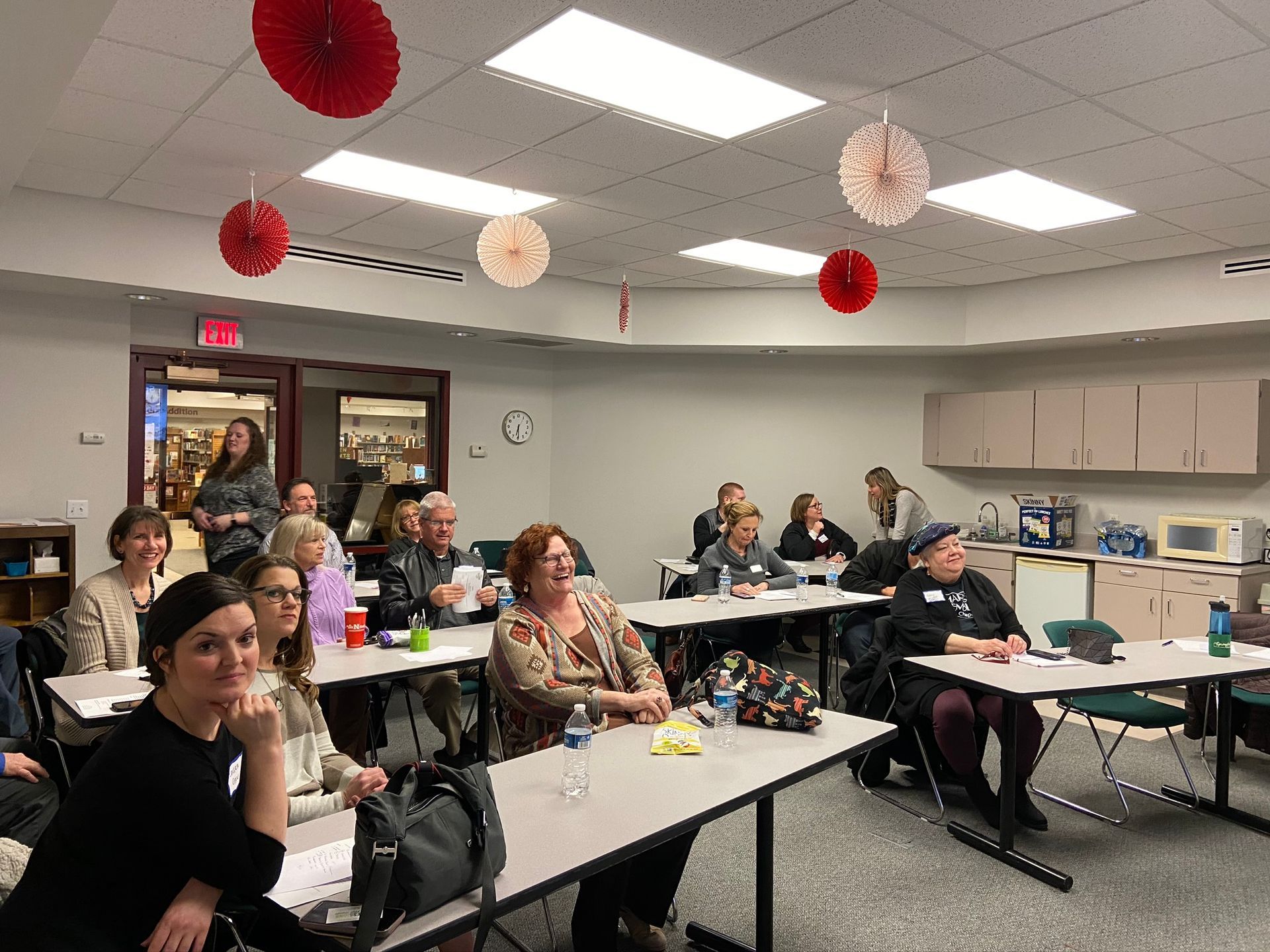 People in a classroom setting, some smiling, with decorative red and white paper fans hanging from the ceiling.