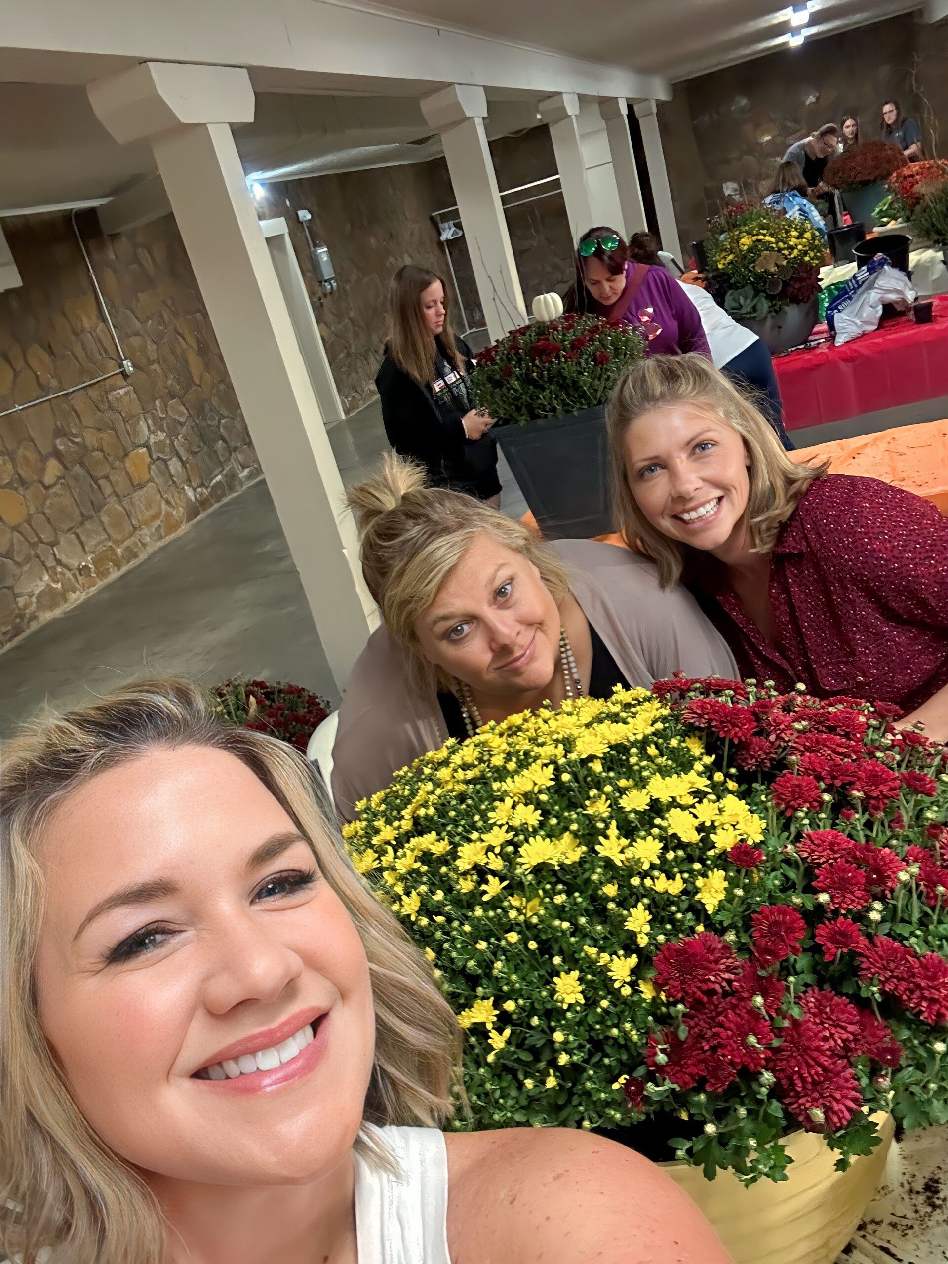 Women smiling, surrounded by colorful potted mums in an indoor space.