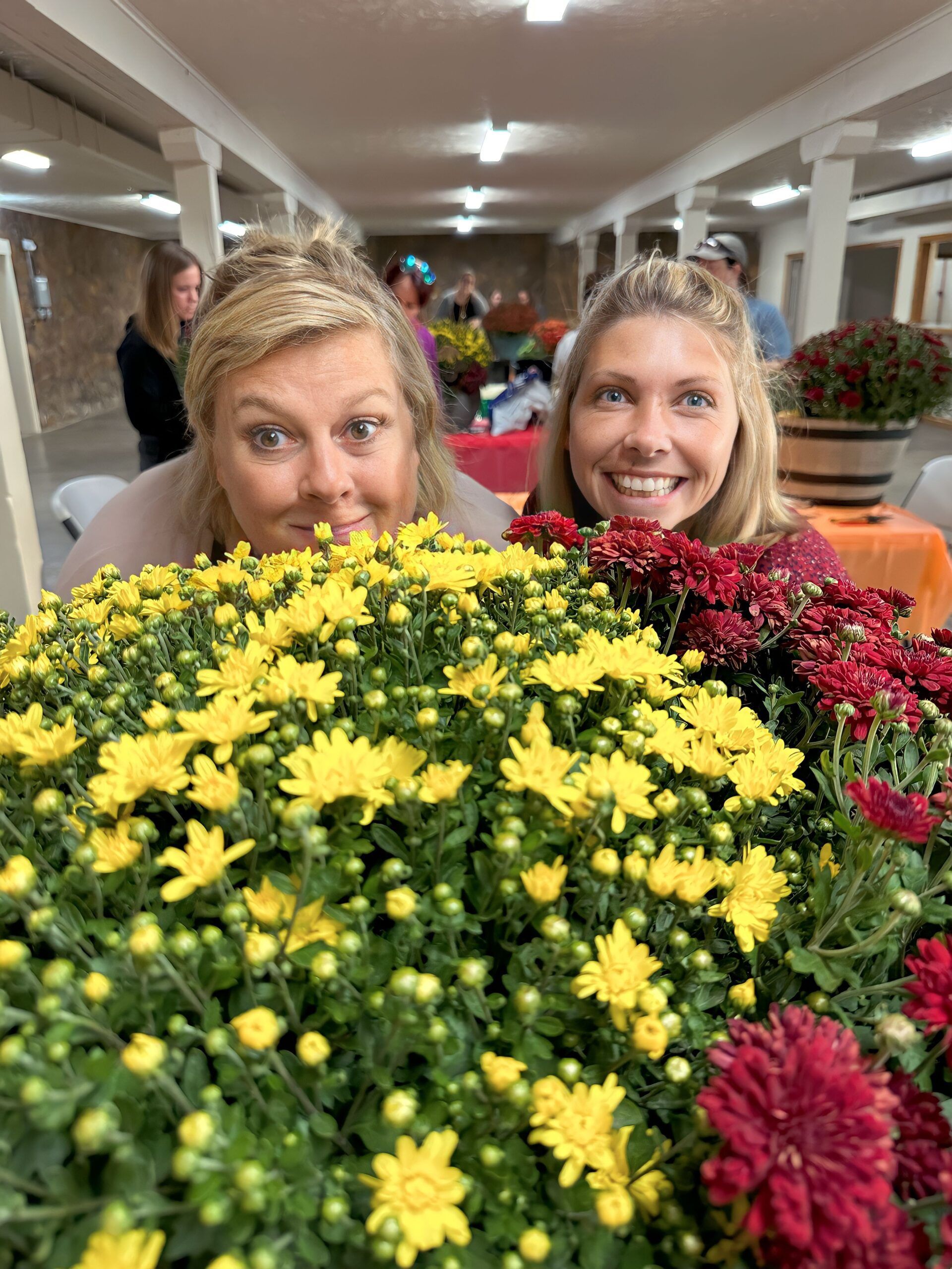 Two women smiling over yellow and red chrysanthemums at an indoor event.
