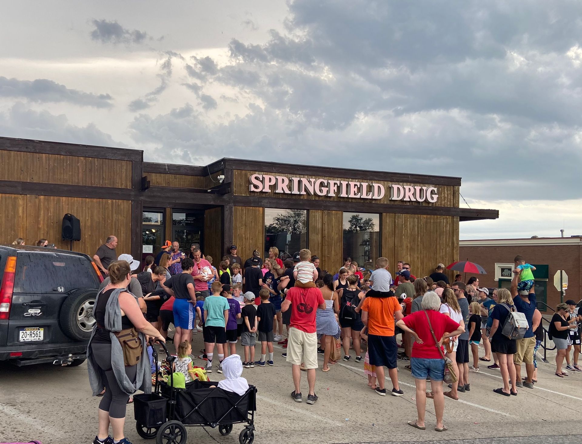 People gathered outside Springfield Drug, a wooden-fronted building. A crowd is visible, possibly attending a