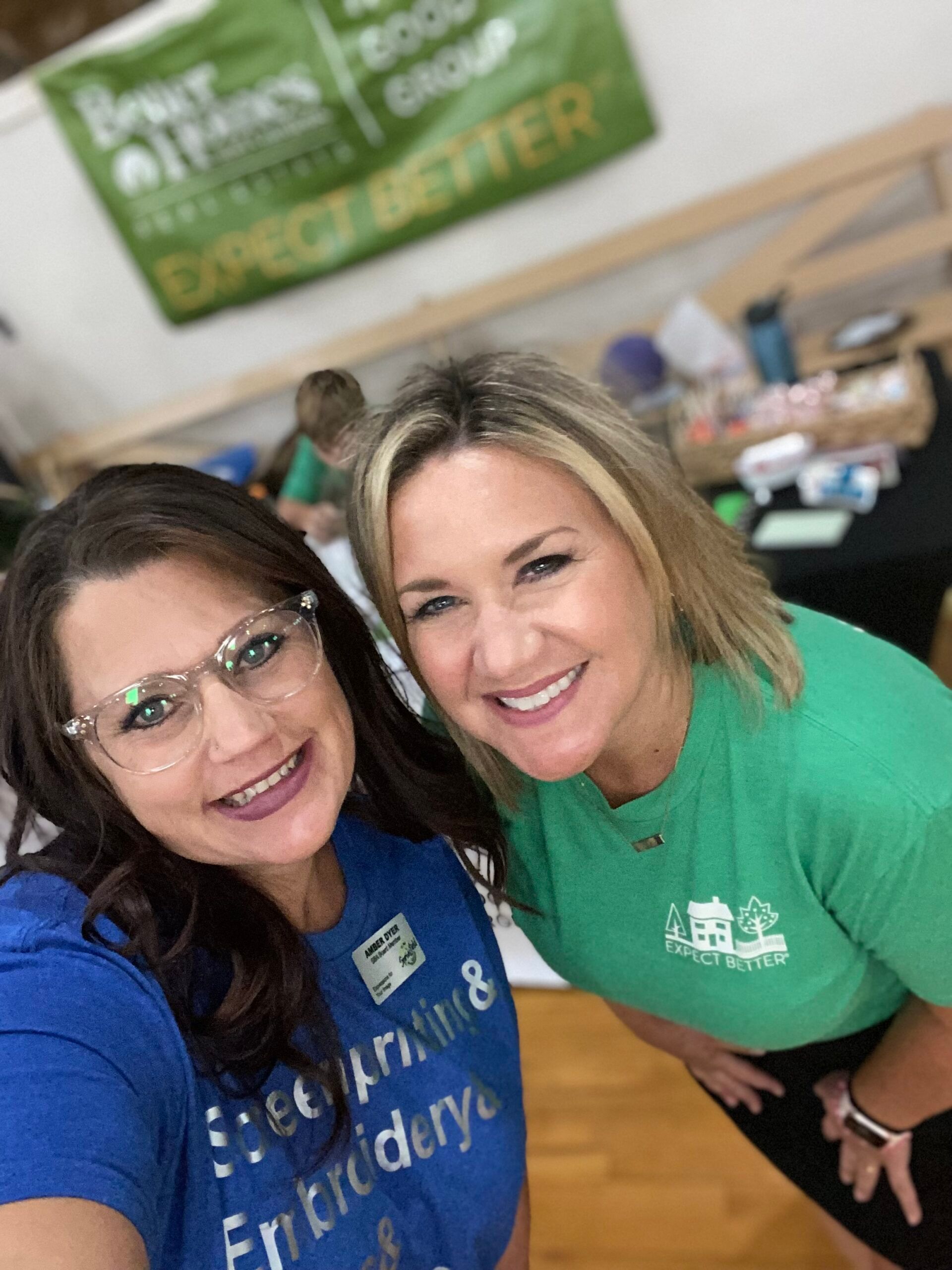 Two smiling women posing for a selfie at an event, one in blue, one in green shirts, and a banner in the background.