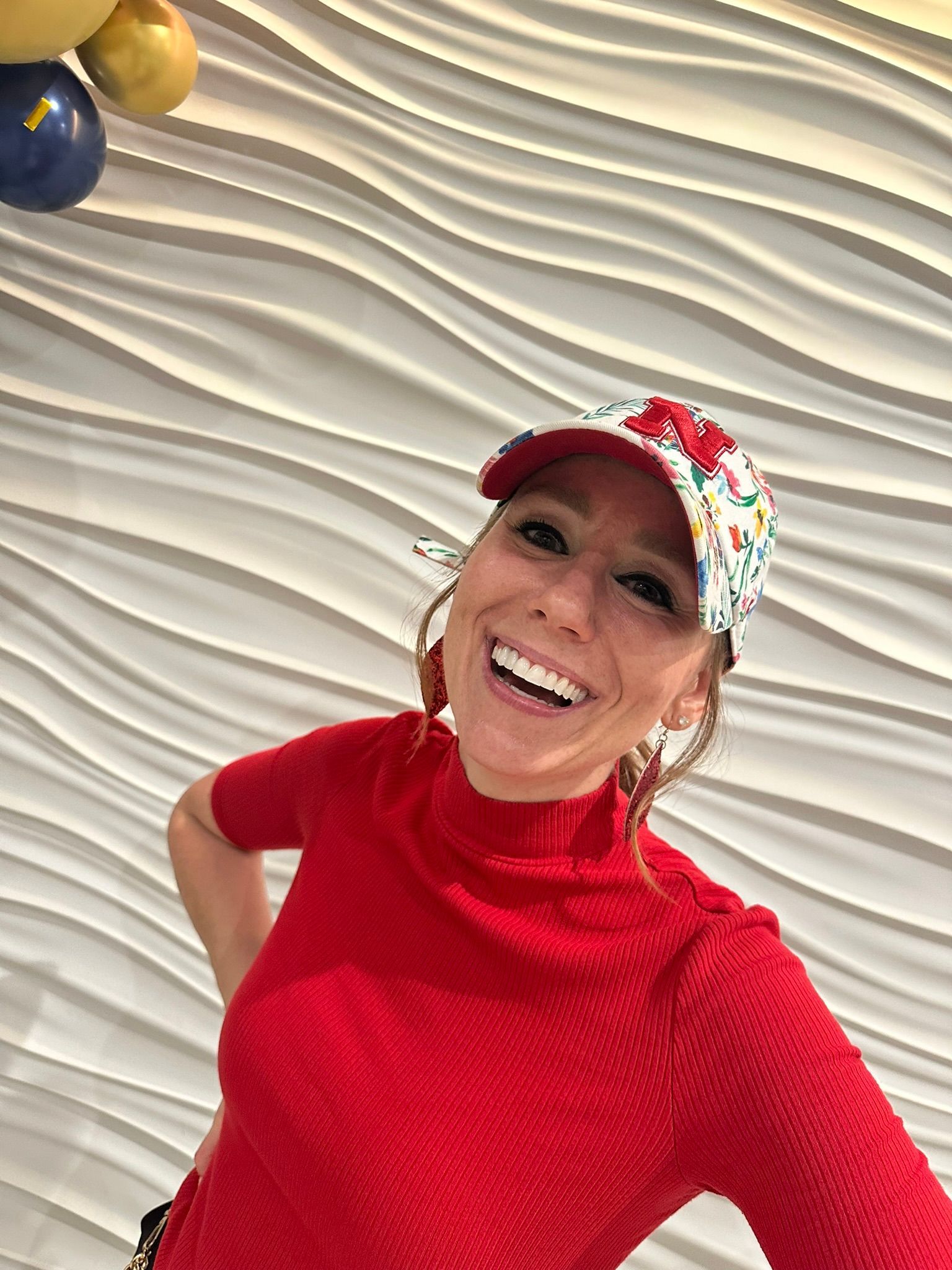 Woman smiling, wearing a floral hat and red shirt, in front of a white wave-patterned wall.