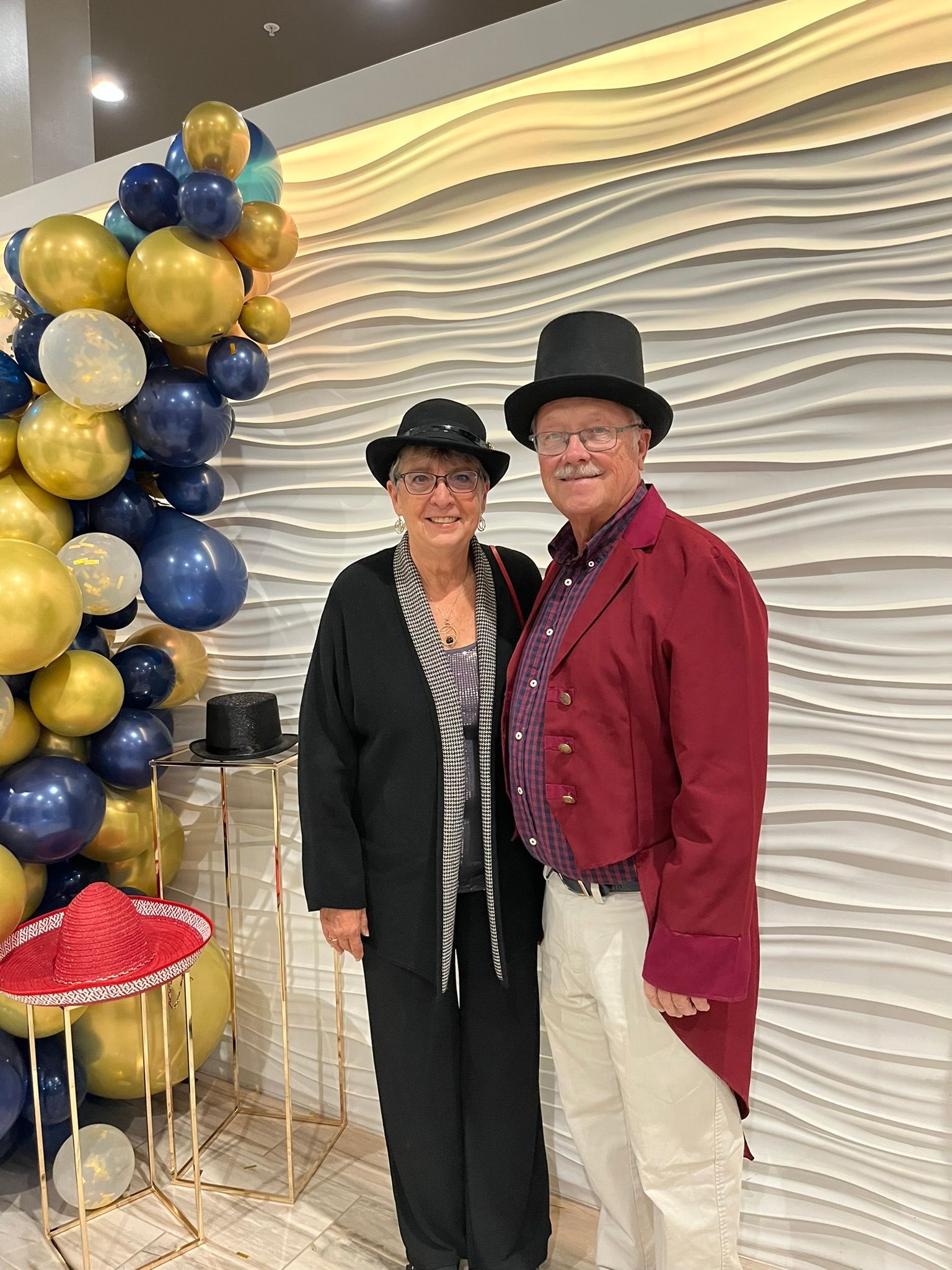 Couple in top hats and festive attire posing near a balloon arch and textured wall.