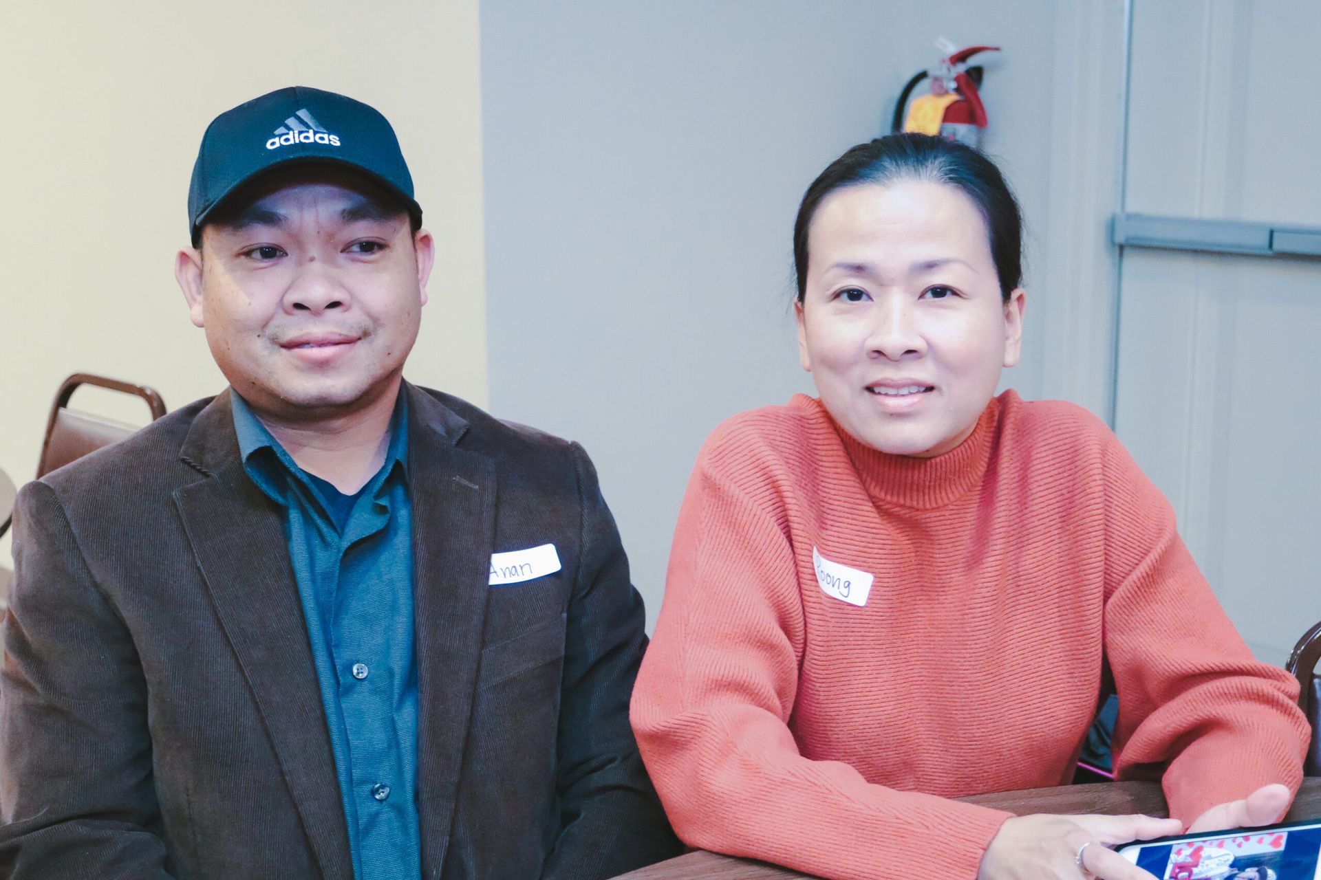 Man in a brown blazer and baseball cap, and woman in an orange sweater, seated and smiling.