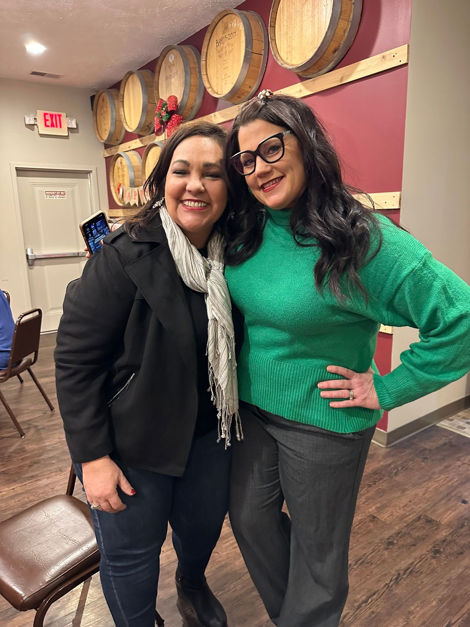 Two women smiling, posing for a photo in front of wine barrels and a burgundy wall. One in a black coat, one in a green sweater.