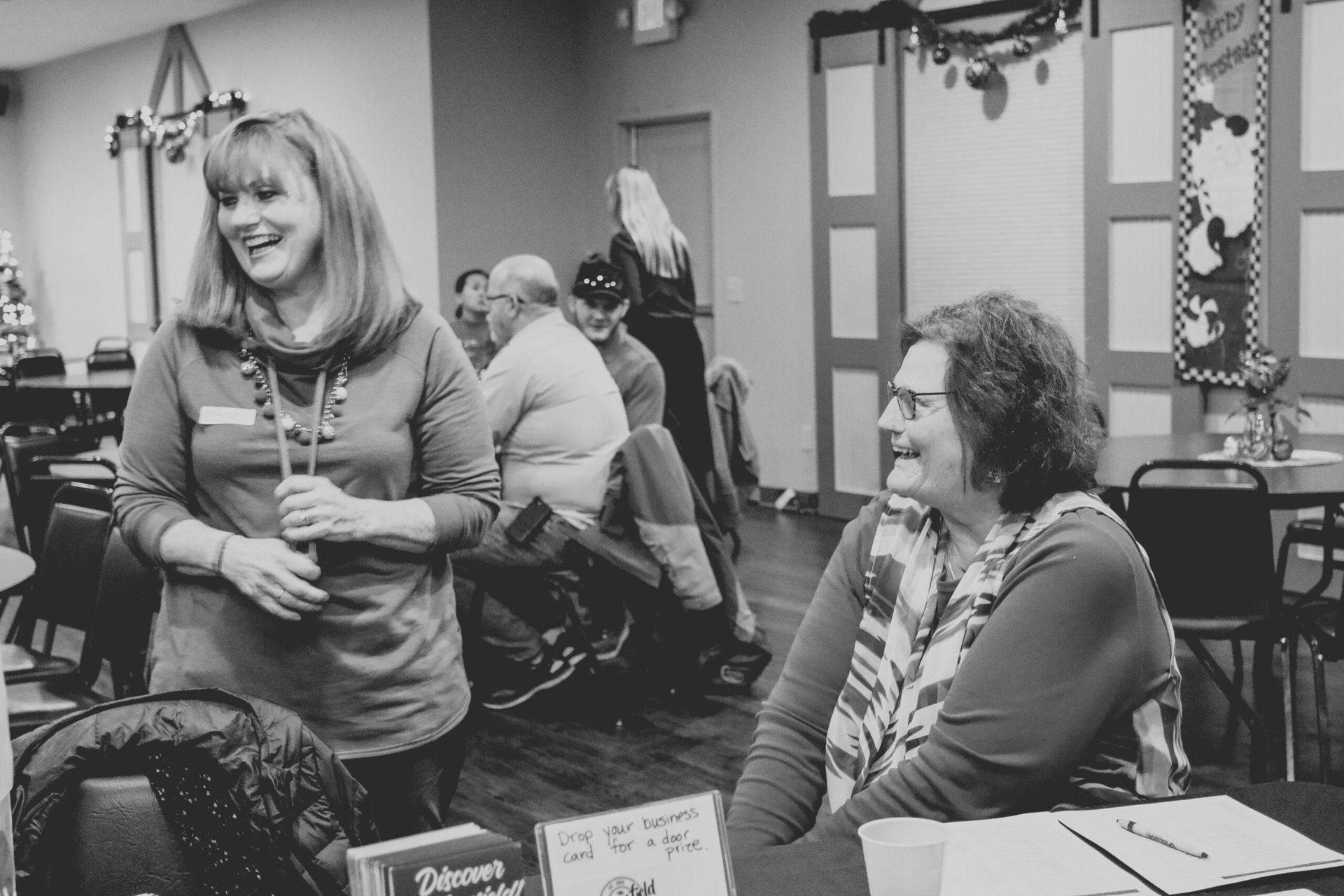 Two women smiling in a room. Others sit at tables and in the background. Festive decorations.