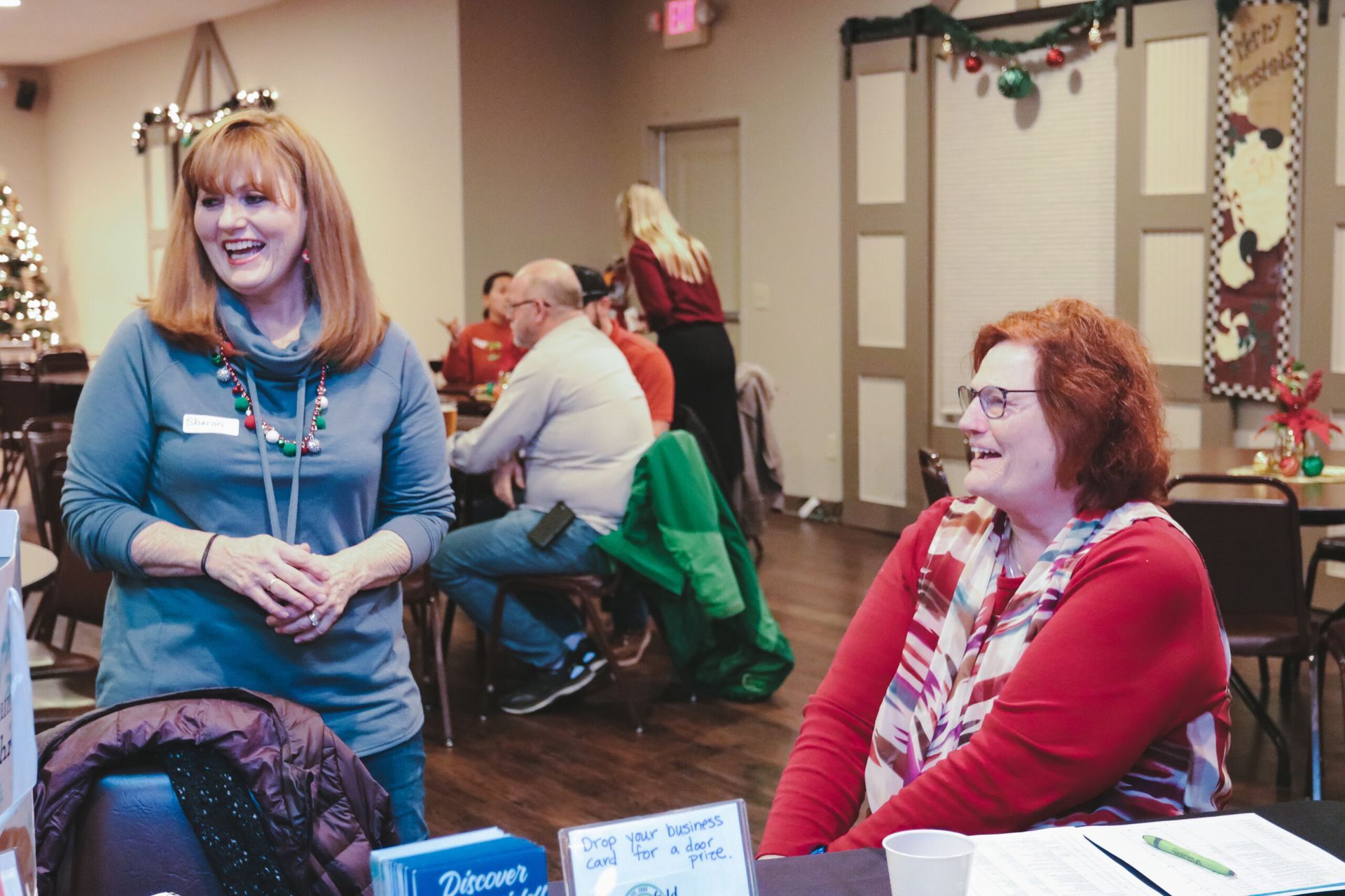 Two women smiling at a table indoors, others in the background at a holiday event.