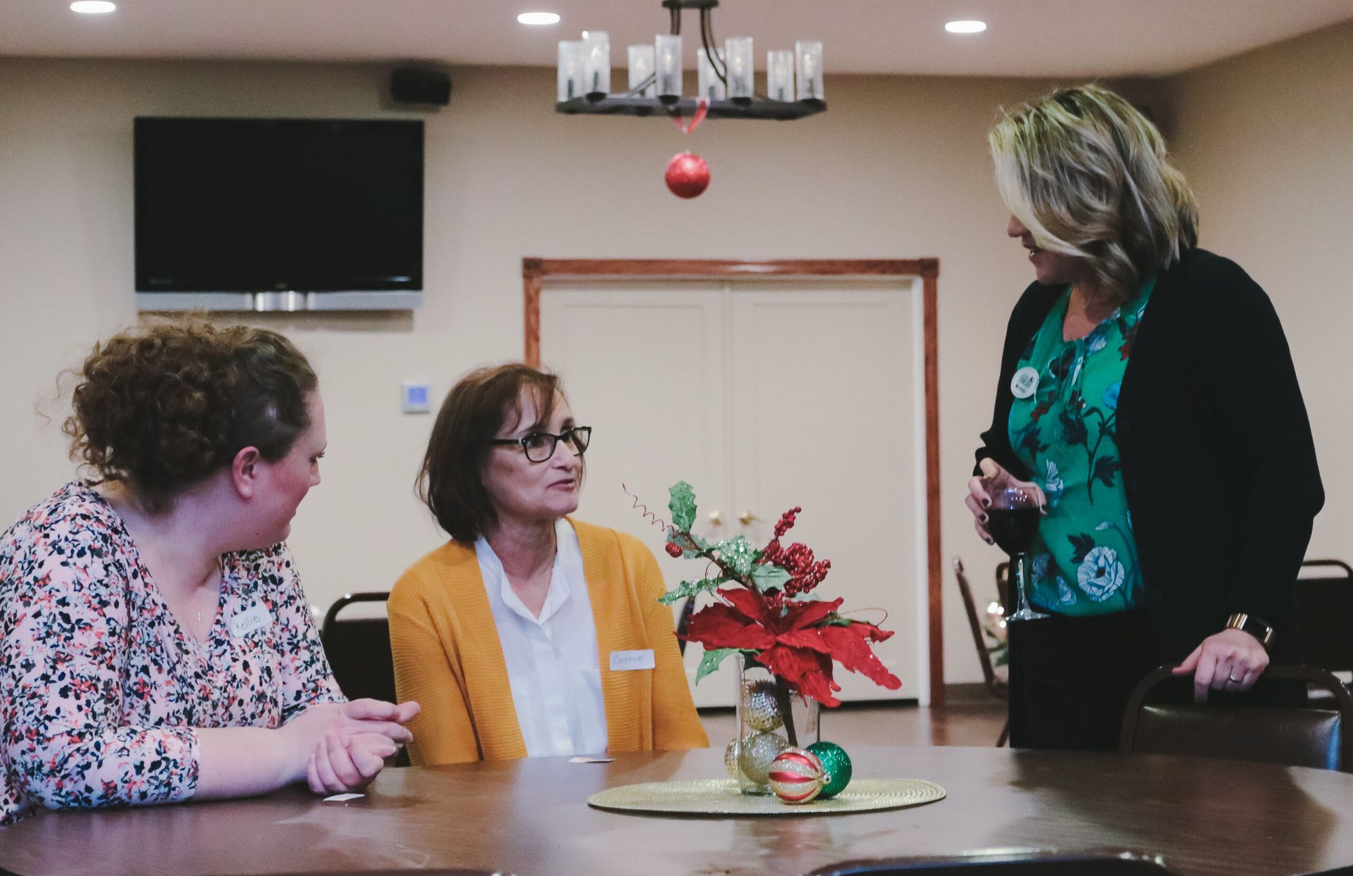 Three women seated at a table, conversing in a decorated room. One woman is gesturing.