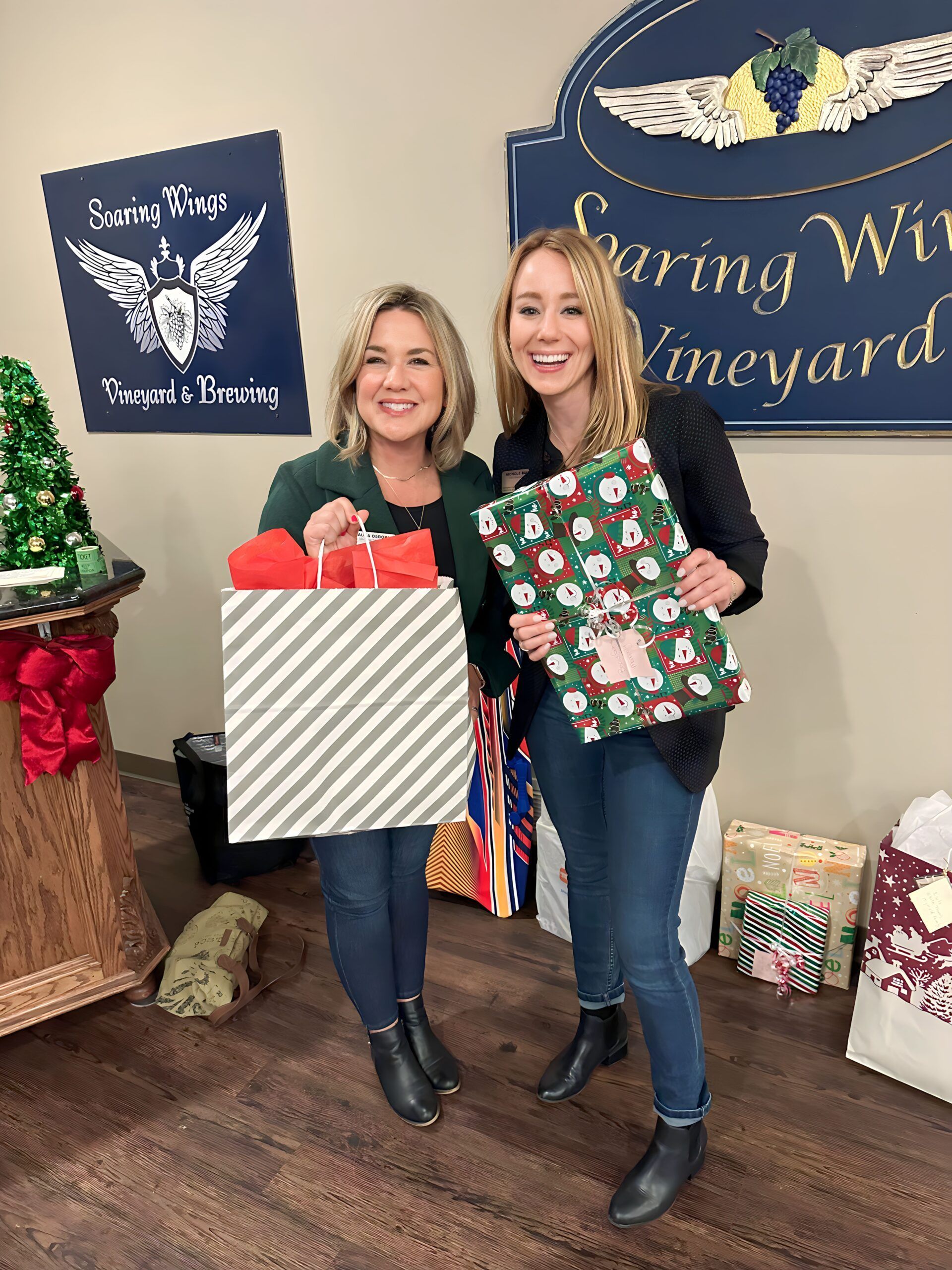 Two women holding wrapped gifts in a vineyard. One with a striped bag, the other with a pig print bag, smiling.