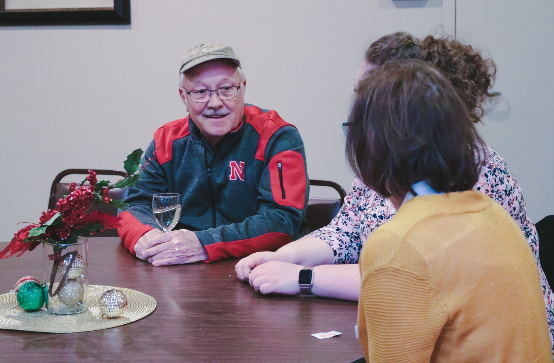 Man in red jacket smiles while holding a glass, seated at table with two others. Festive decor present.